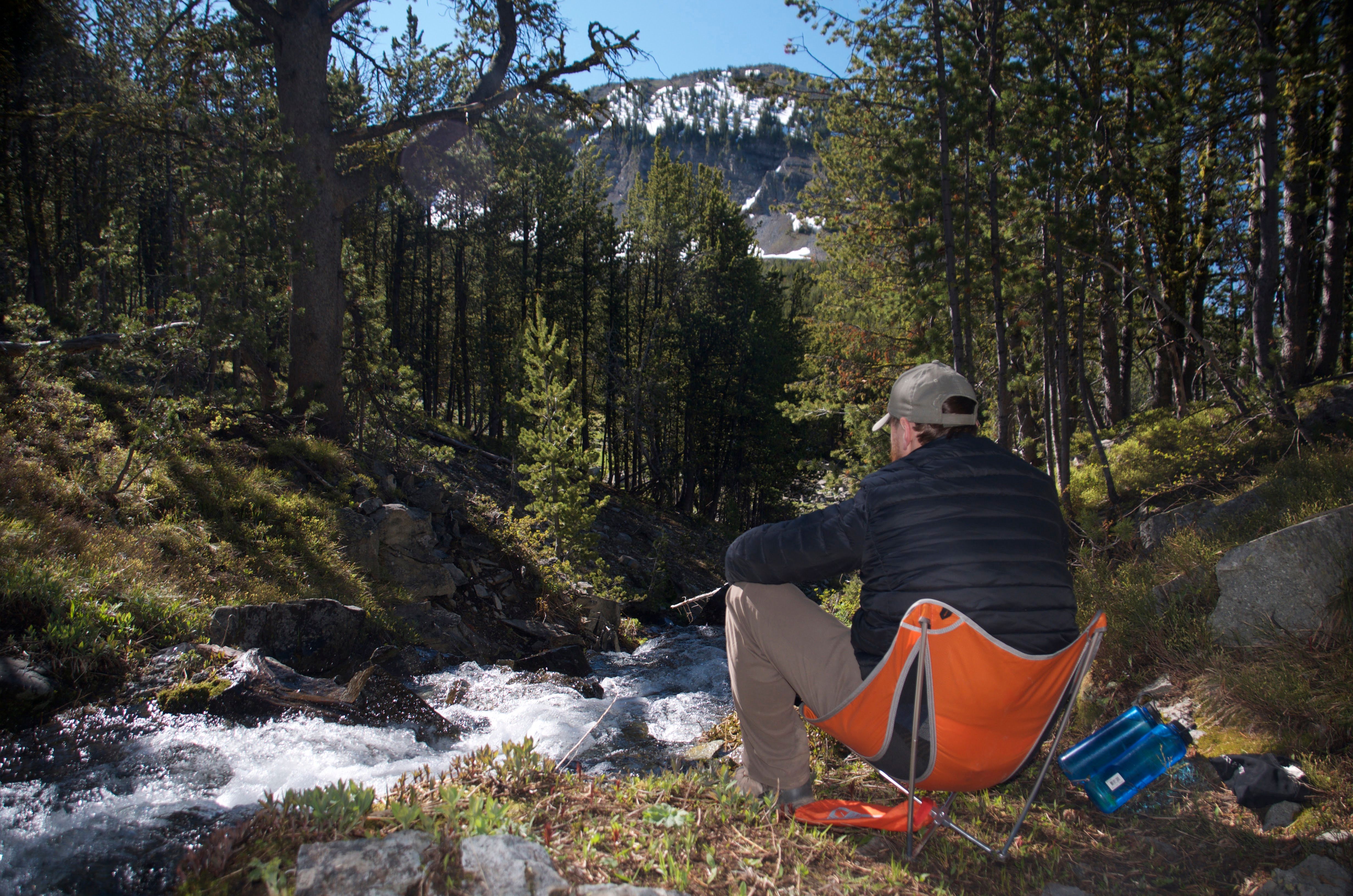 Camper relaxing in a chair beside a rushing mountain creek with snow-capped peaks in the background