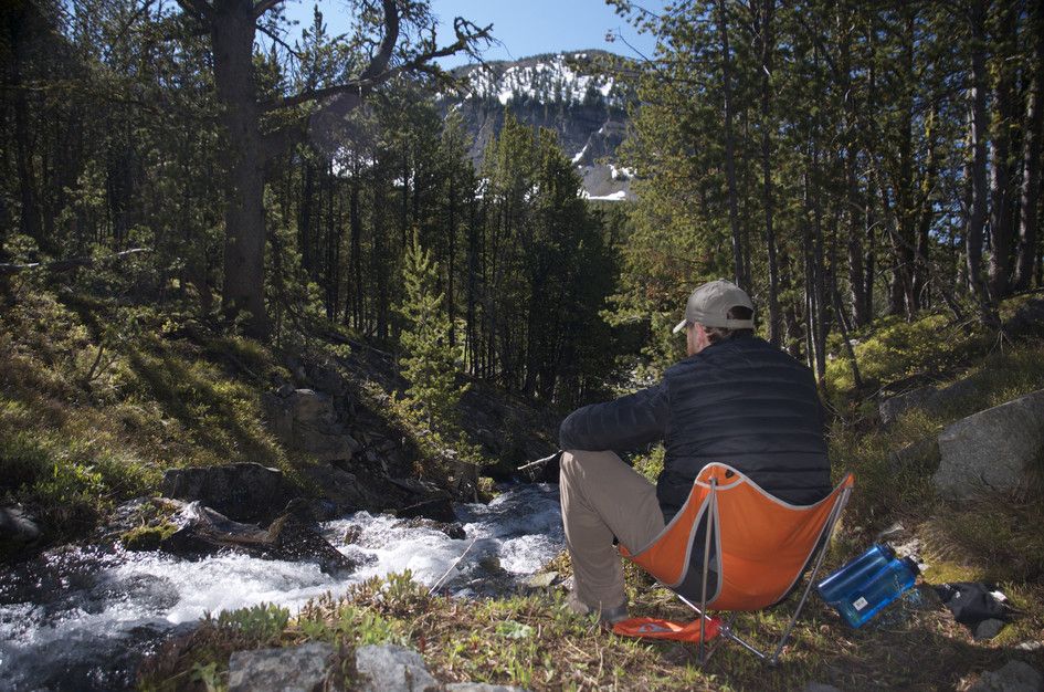 Hiker sitting in camp chair by rushing creek with snow-patched mountain