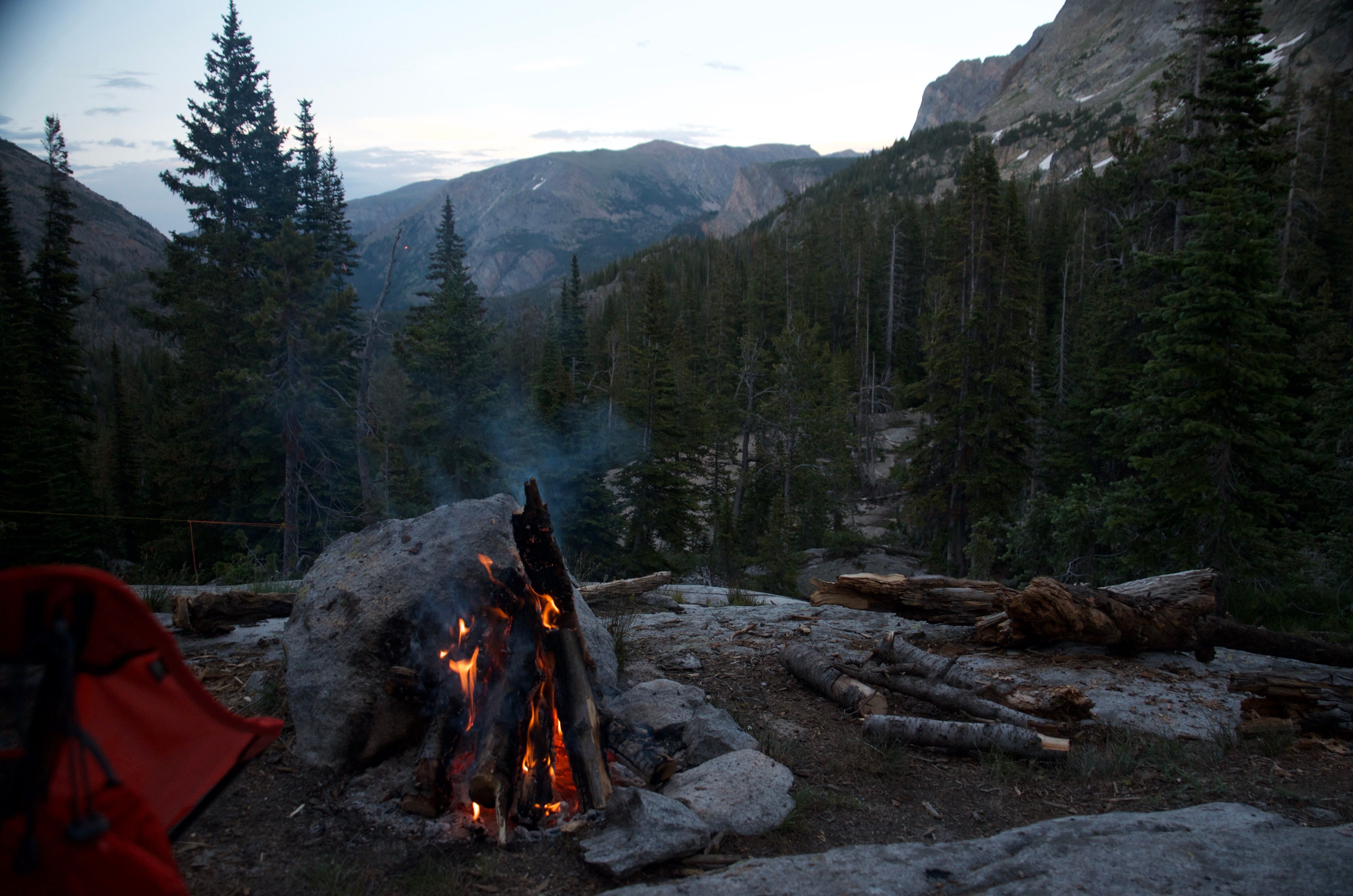Campfire in a rock fire ring at dusk with mountain peaks and evergreen forest