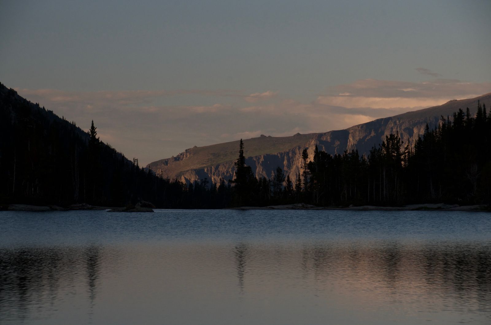 Peaceful alpine lake at dusk with silhouetted pines and distant cliffs