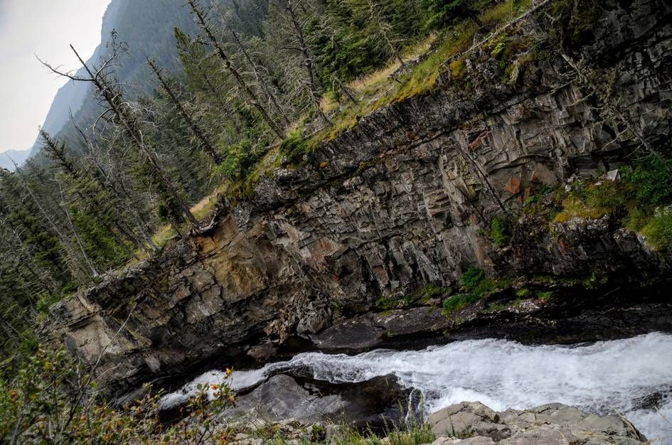 Layered rock cliff with rushing waterfall and sparse forest on steep slope