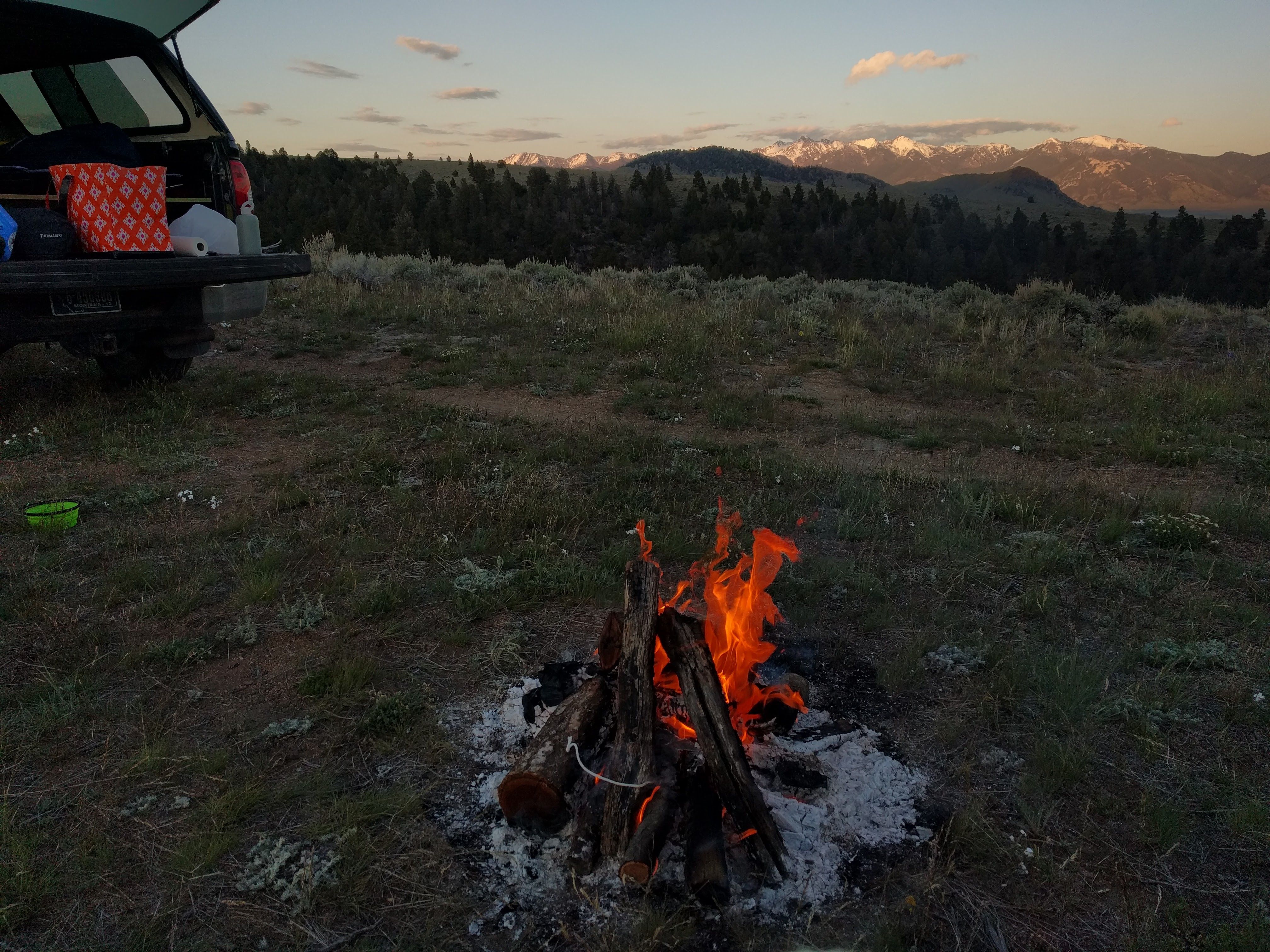 Campfire at sunset with snow-capped mountains visible behind the treeline