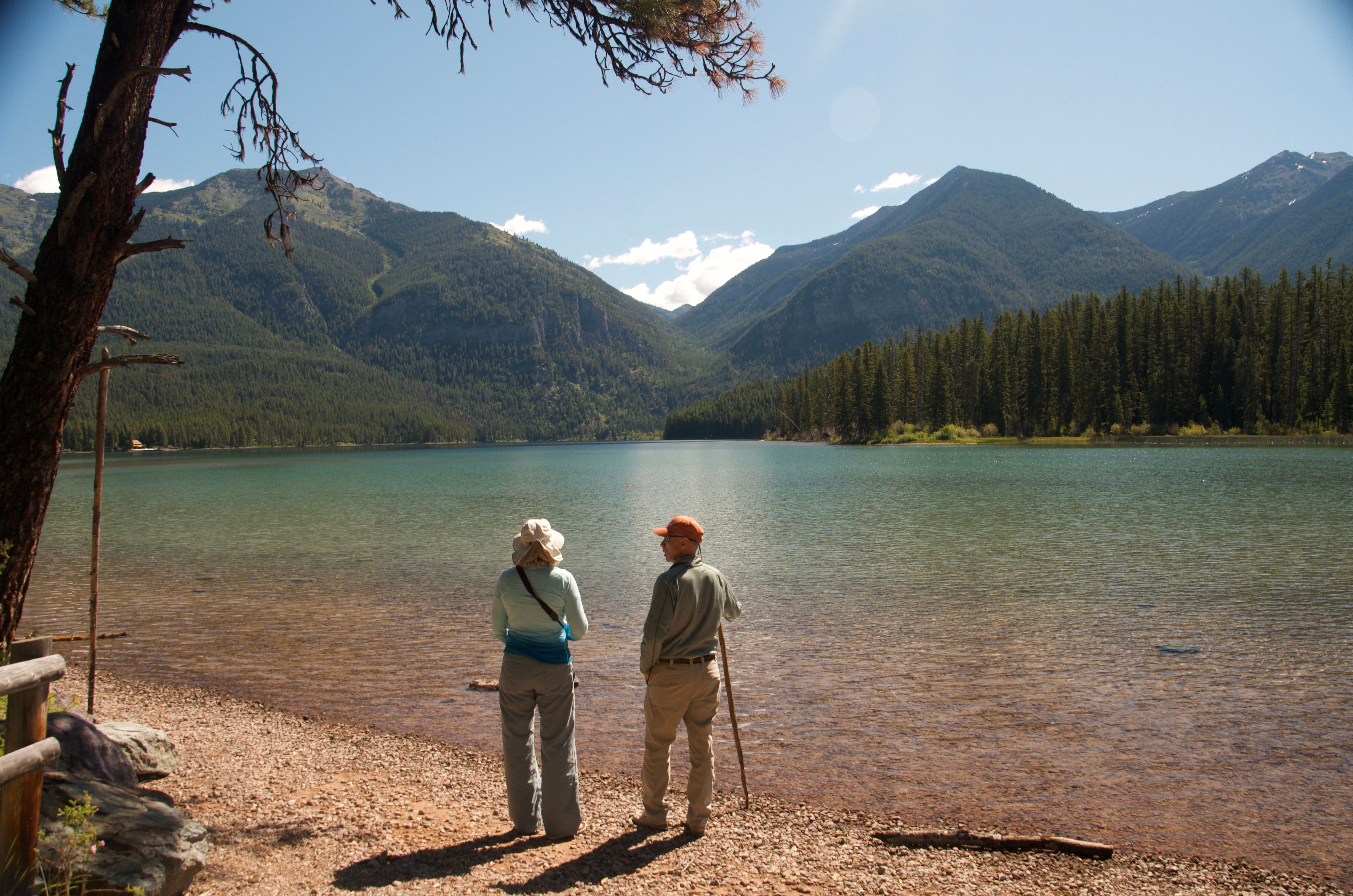 Two hikers standing at the edge of Holland Lake looking toward snow-capped mountains
