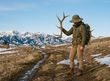 Hiker holding up a set of elk antlers on a dry hillside trail with snow-capped mountains in the background