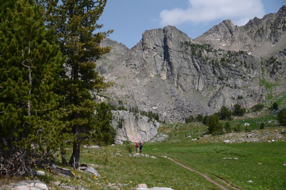 Hikers on trail through green meadow dwarfed by towering granite cliffs