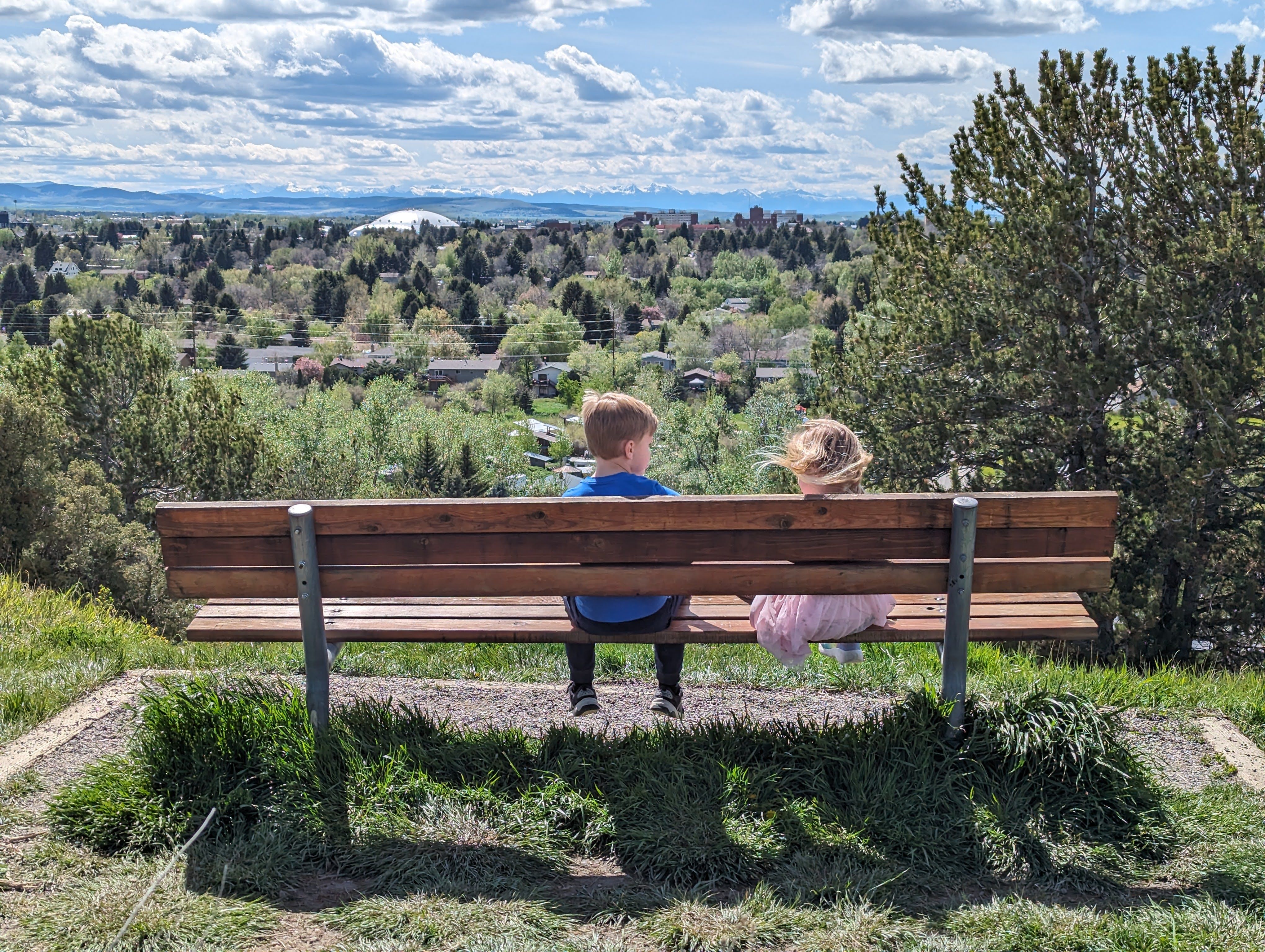 Two young children sitting on a bench at a Burke Park overlook with their backs to the camera, looking out over Bozeman in spring