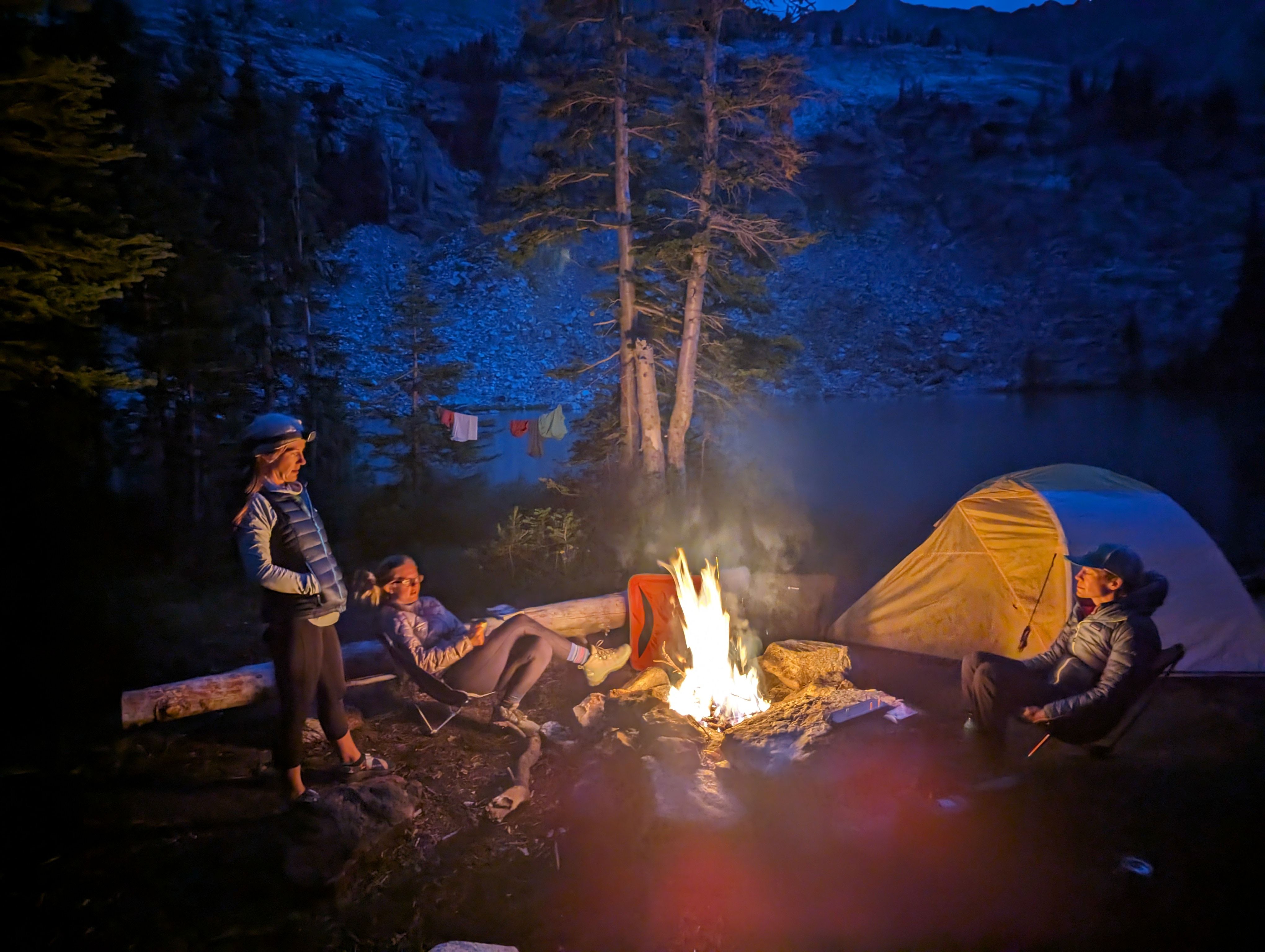 Three friends around a campfire at an alpine lake at night with a tent and rocky peaks behind them