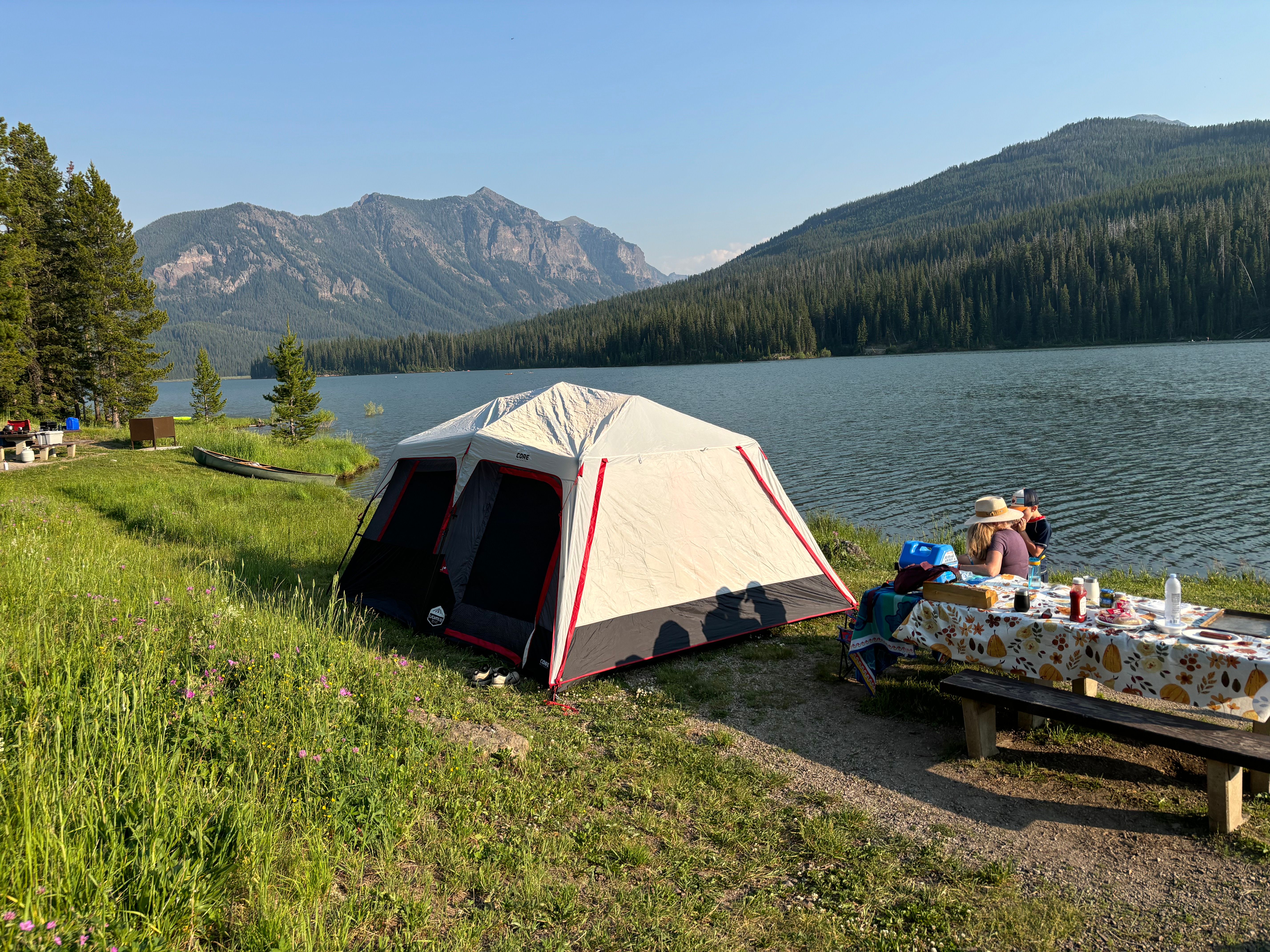 Tent pitched on a grassy lakeshore with mountains and pine forest behind, picnic table with campers in the foreground