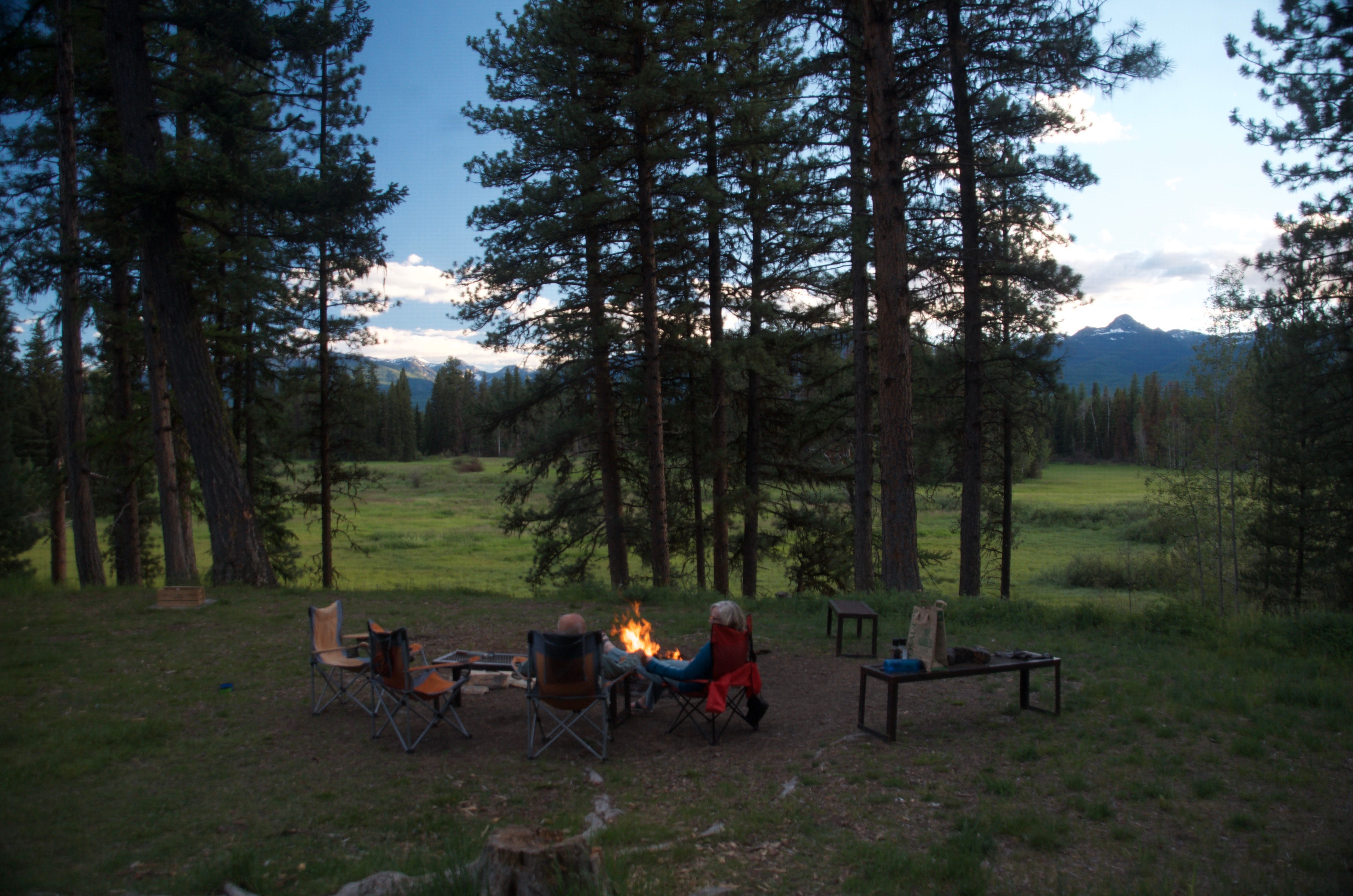 Small group sitting around a campfire at dusk with the open meadow and distant mountains behind them
