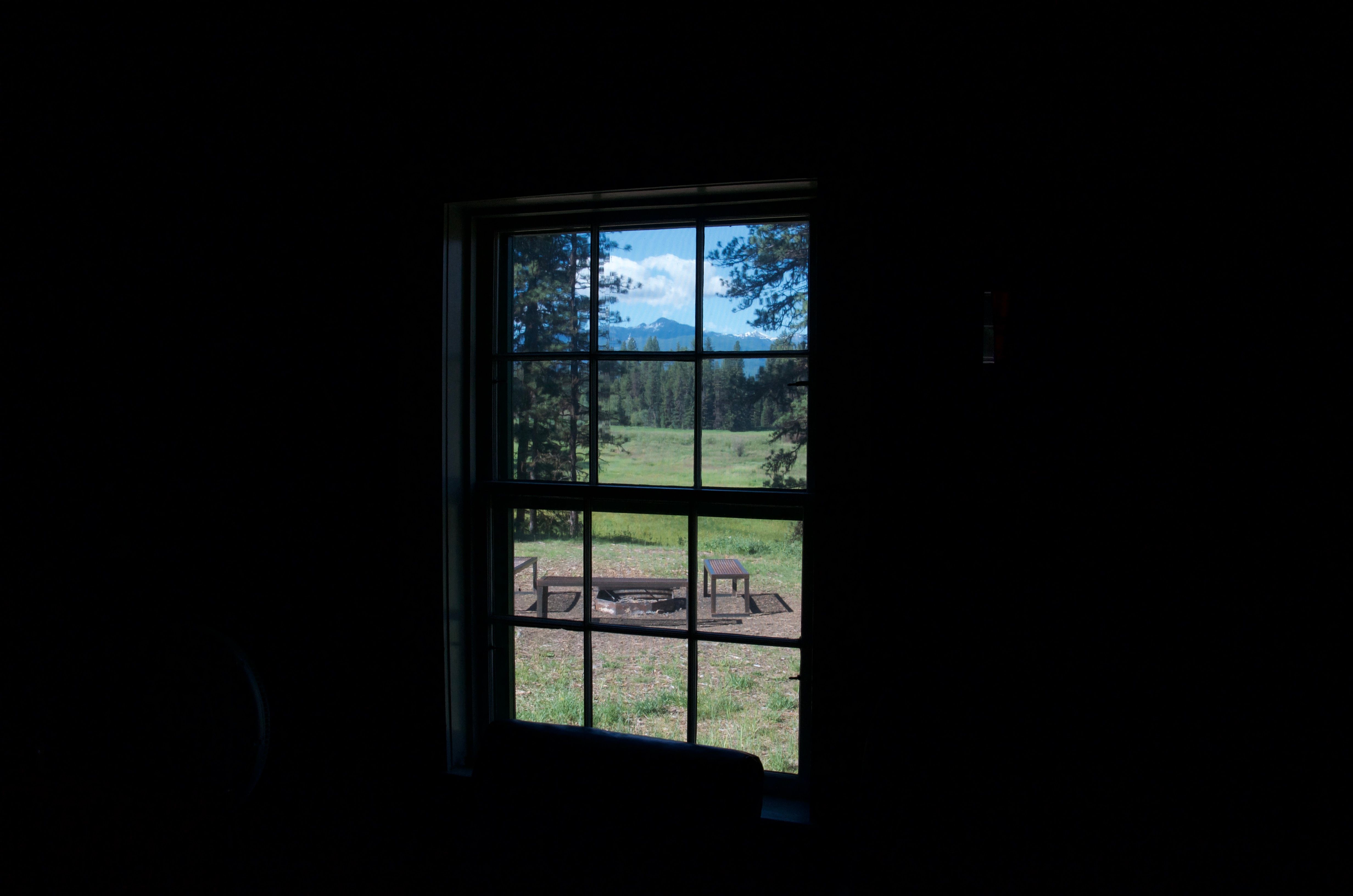 View through a cabin window of the fire pit area and the open meadow with snow-capped mountains in the distance
