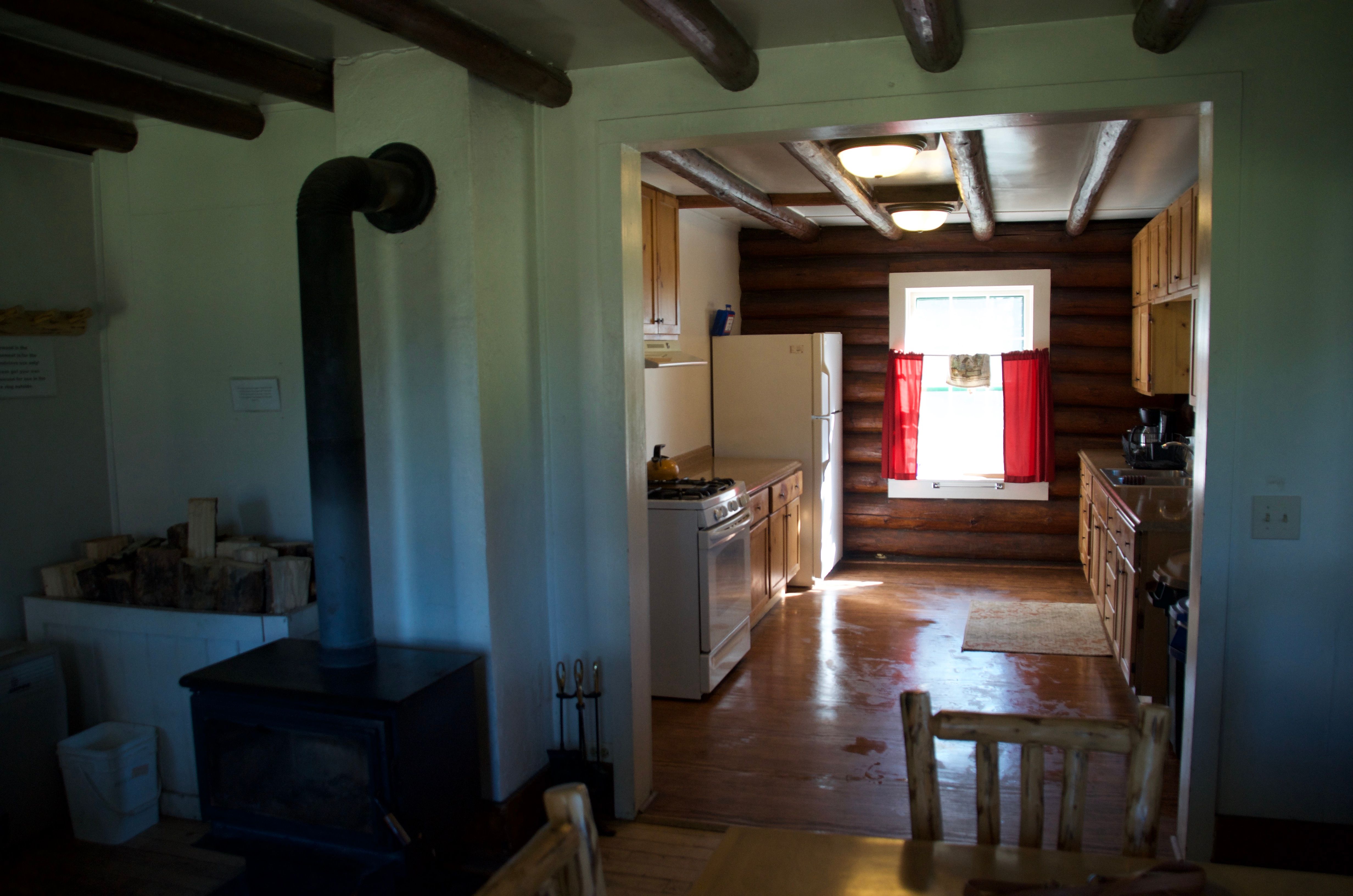 Wood burning stove with stacked firewood in the foreground and the kitchen visible through the doorway
