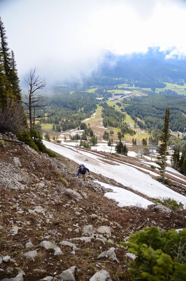 Hiker scrambling up steep rocky slope with ski area and valley far below