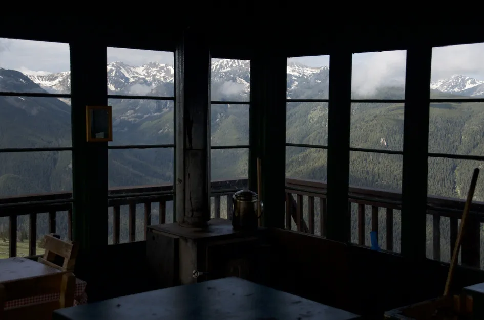 View from inside the Garnet Mountain Fire Lookout, looking out through wraparound windows at snow-capped peaks and forested valleys