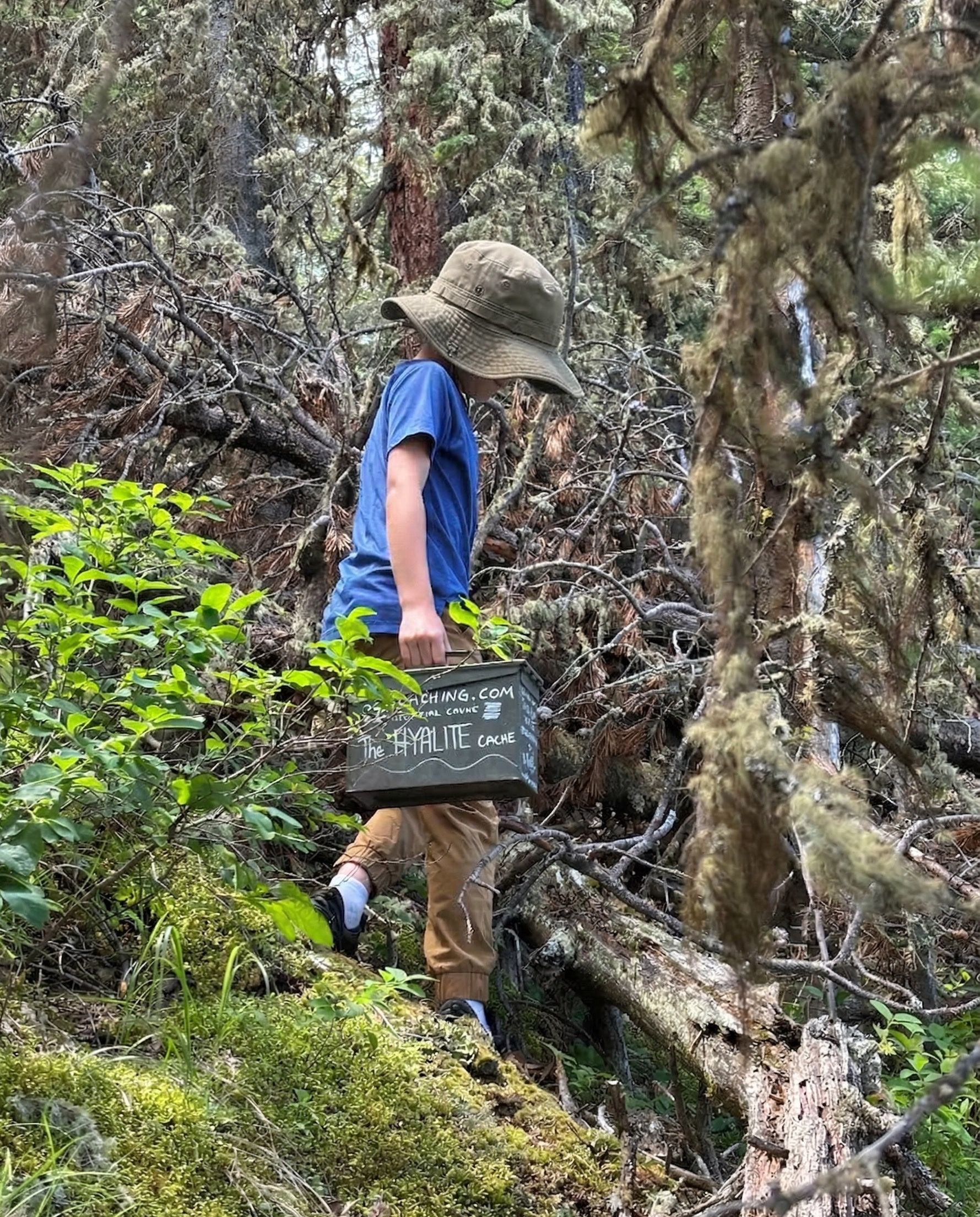 Boy in bucket hat holding the Hyalite Cache geocache box in the woods