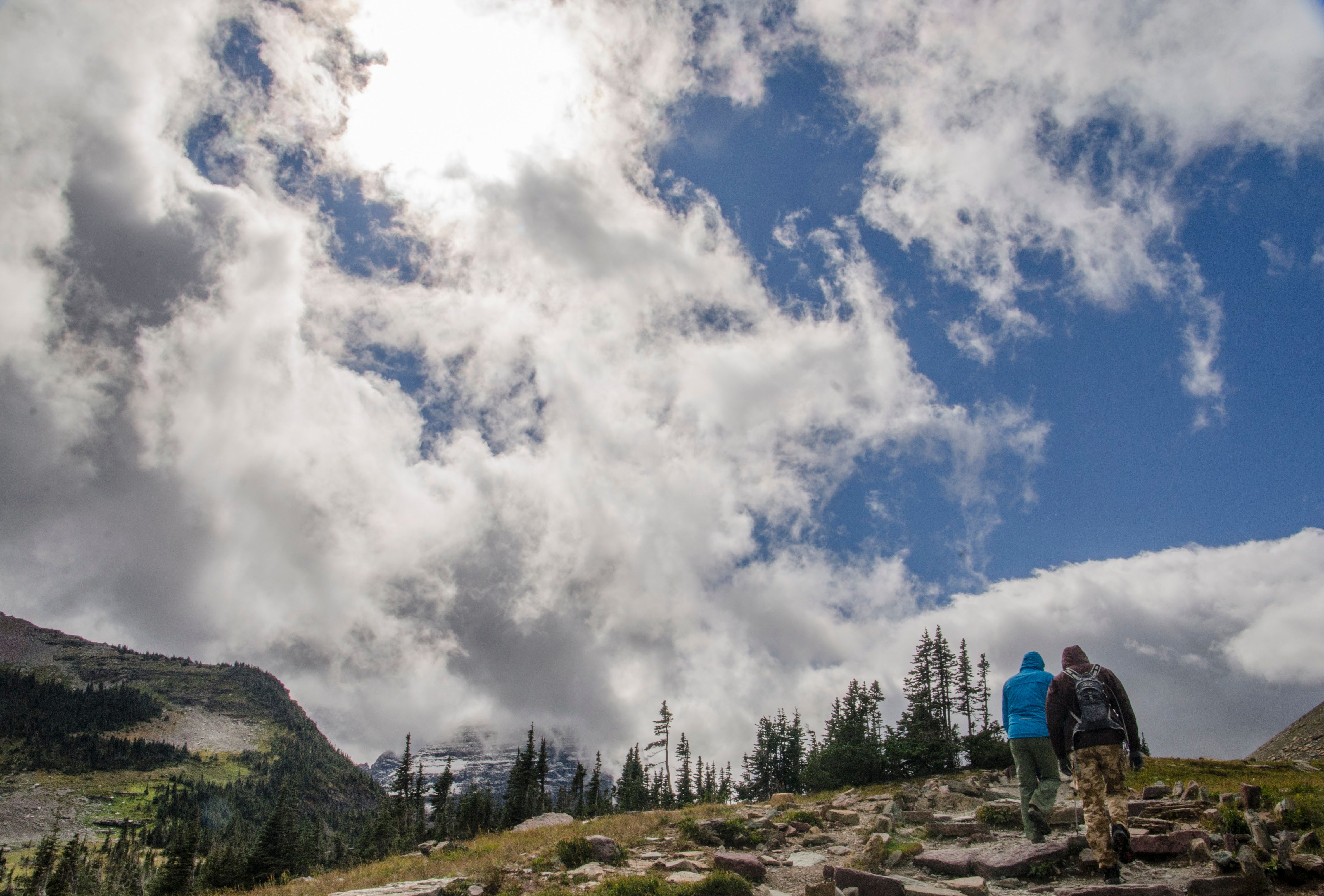 Two hikers walking on rocky trail toward mountains beneath towering clouds