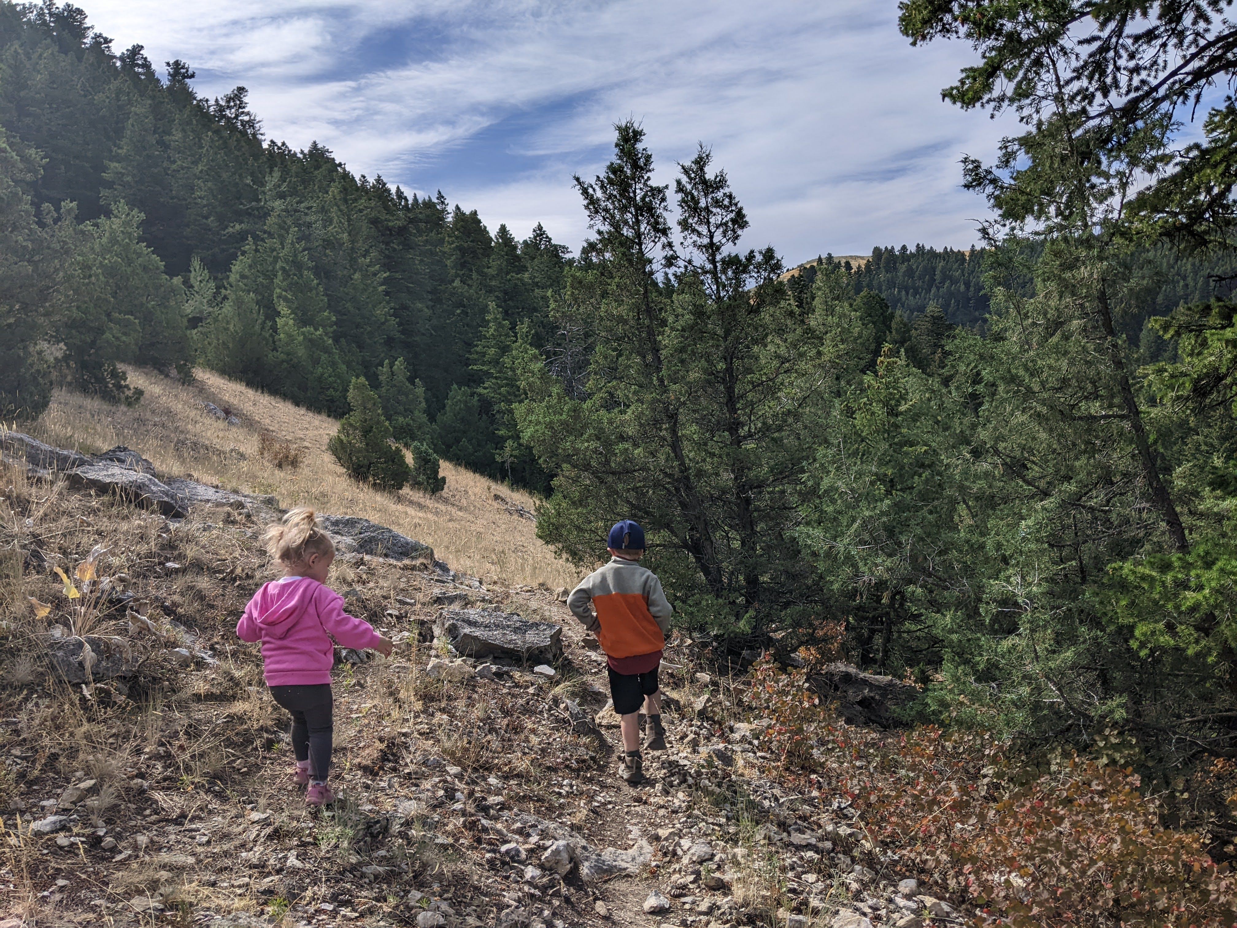 Two kids hiking up a rocky open hillside trail toward the M with pine forest and valley views behind them