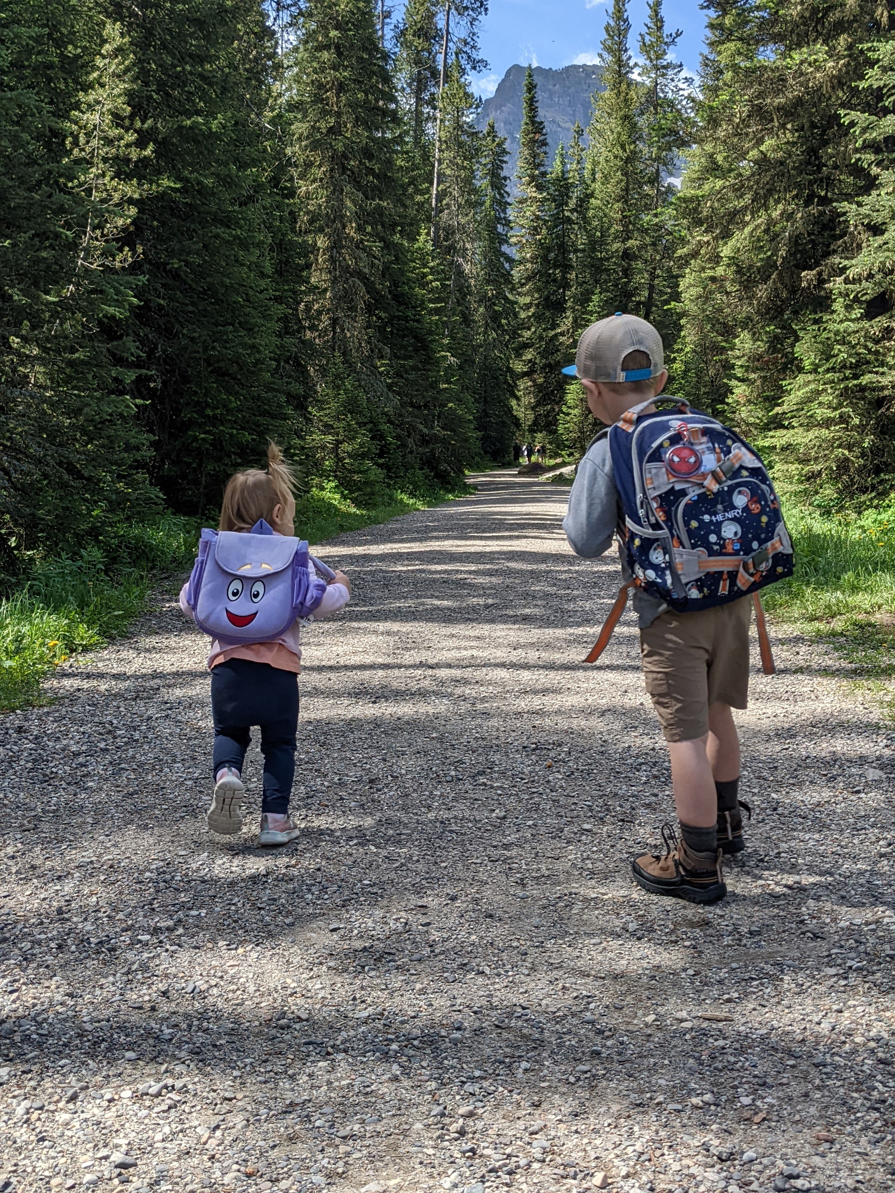 Two young children with small backpacks walking down a wide gravel trail through pine forest with mountains in the background