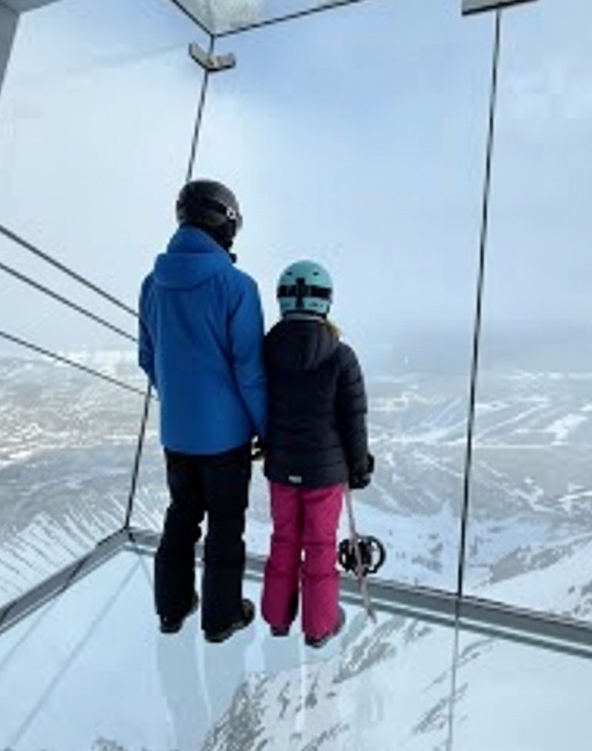 Two visitors standing on the glass floor inside Kircliff observation deck looking out at snow-covered mountains