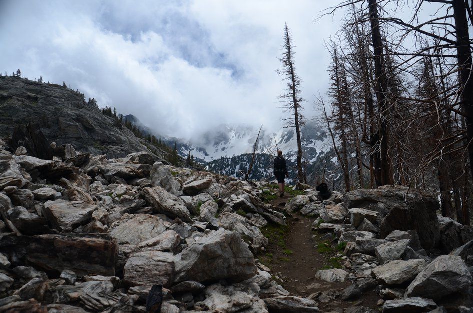 Hikers on rocky trail through boulder field with dramatic cloudy peaks ahead