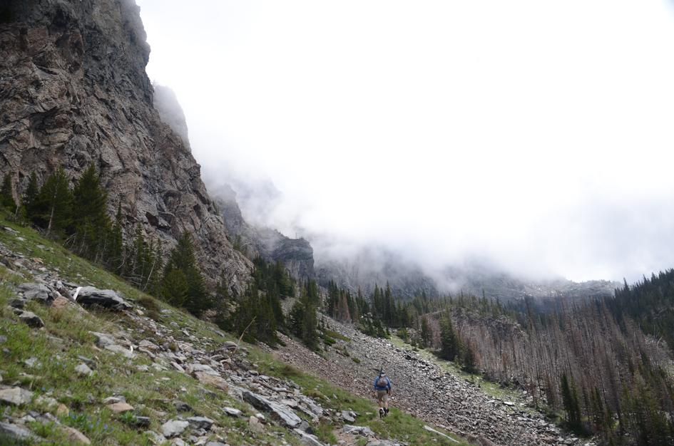 Hiker ascending rocky trail below massive cliff walls with swirling clouds