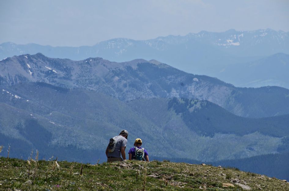 Two hikers sitting on alpine grass enjoying view of layered mountain ridges