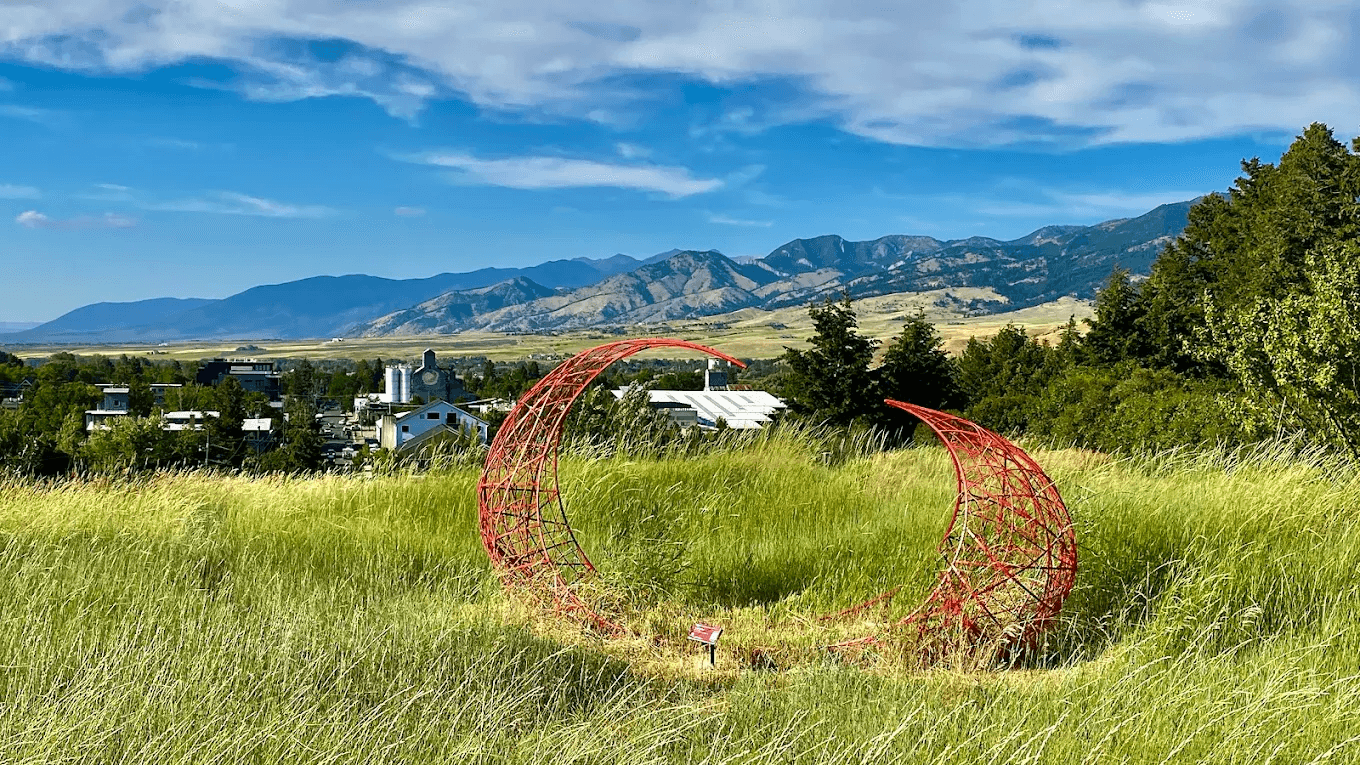 Red circular ring sculpture in green summer grass on the Burke Park hillside with Bozeman and mountains visible behind it