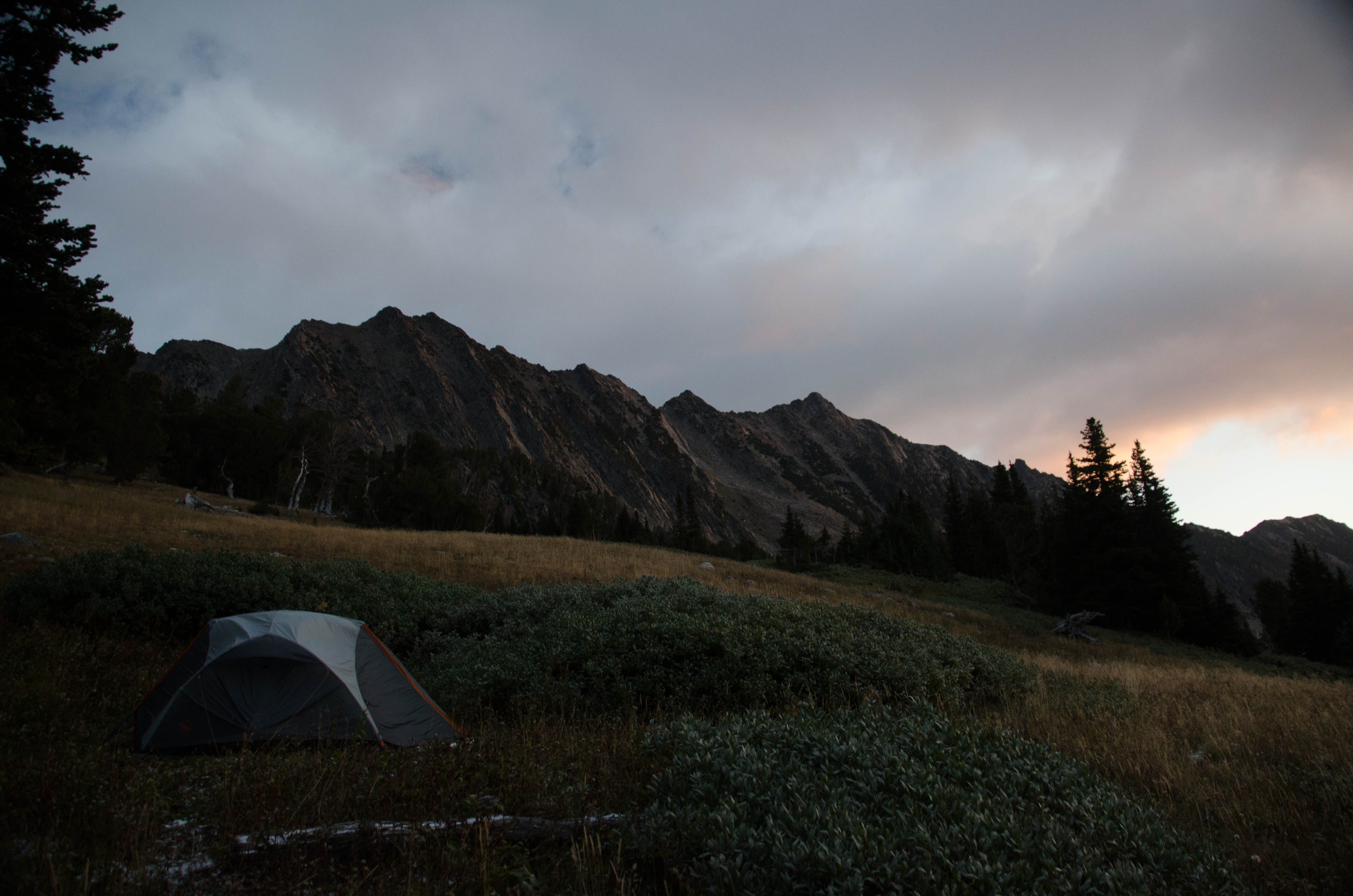 Tent pitched in an alpine meadow below dramatic mountain peaks at dusk