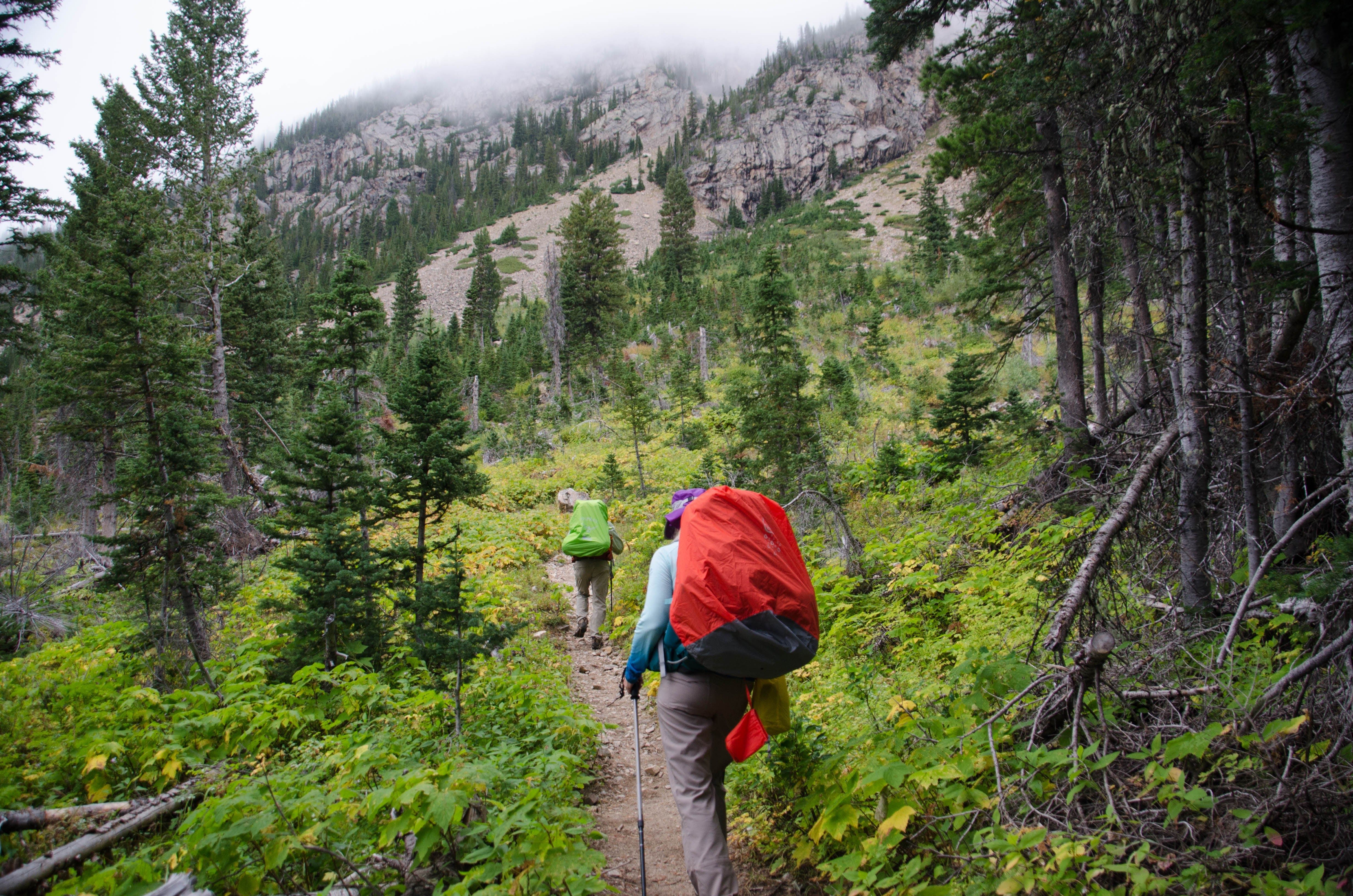 Two backpackers hiking up trail toward rocky peaks with low clouds