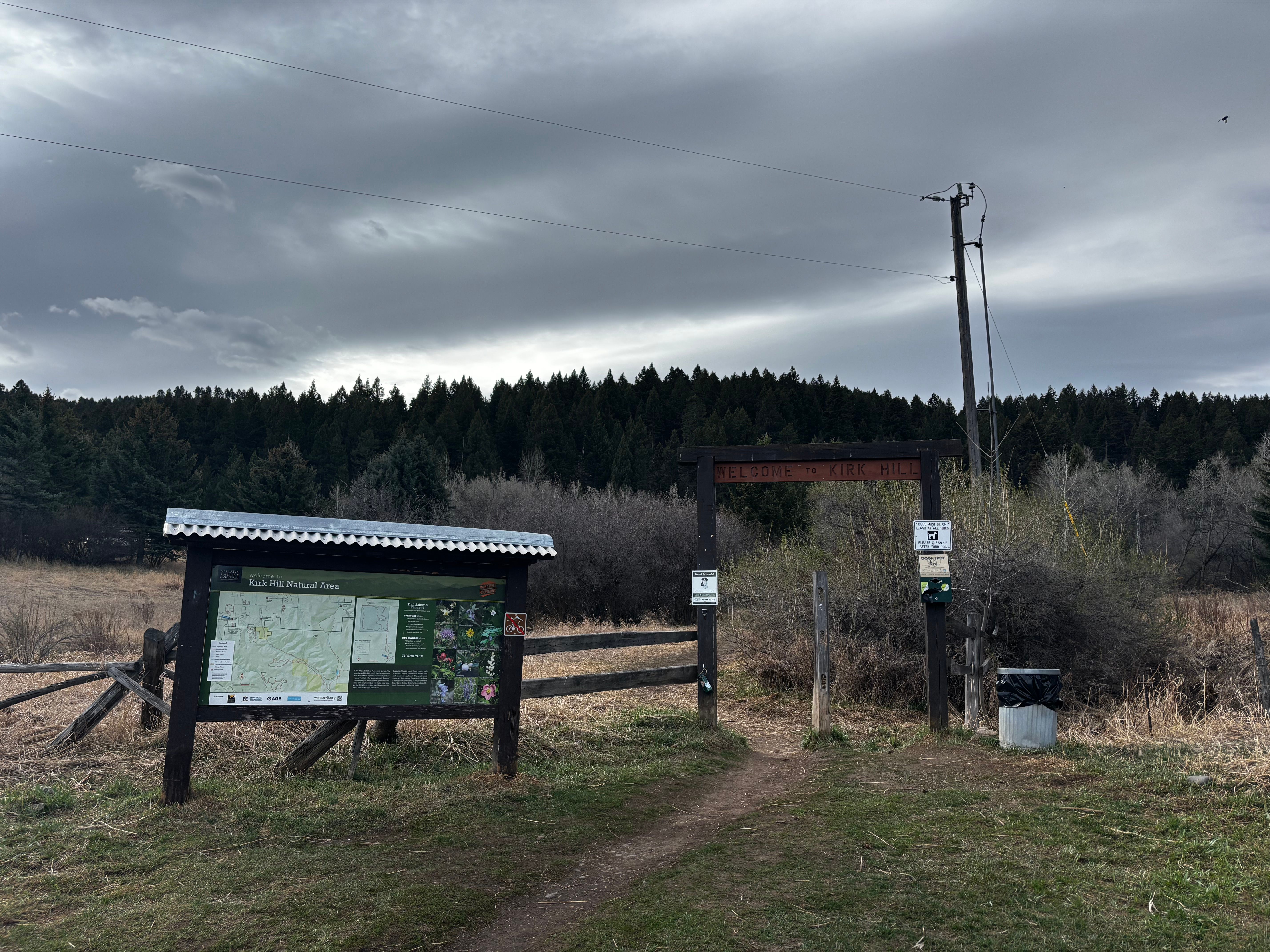 Kirk Hill Natural Area trailhead with information kiosk and Welcome gate under a dramatic cloudy sky