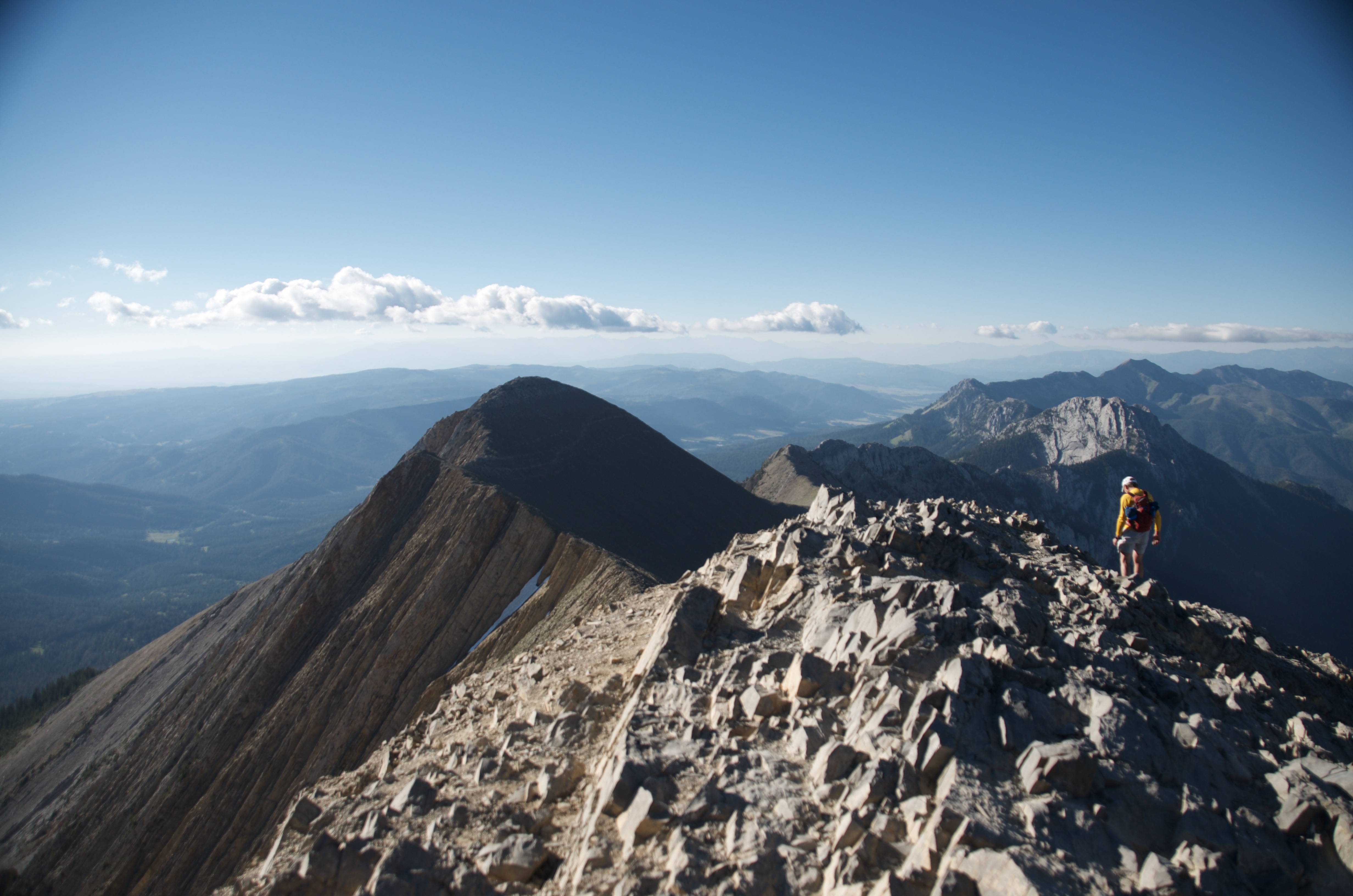 Hiker in yellow jacket standing on narrow rocky Bridger Ridge spine with dramatic shadow peak and Gallatin Valley far below