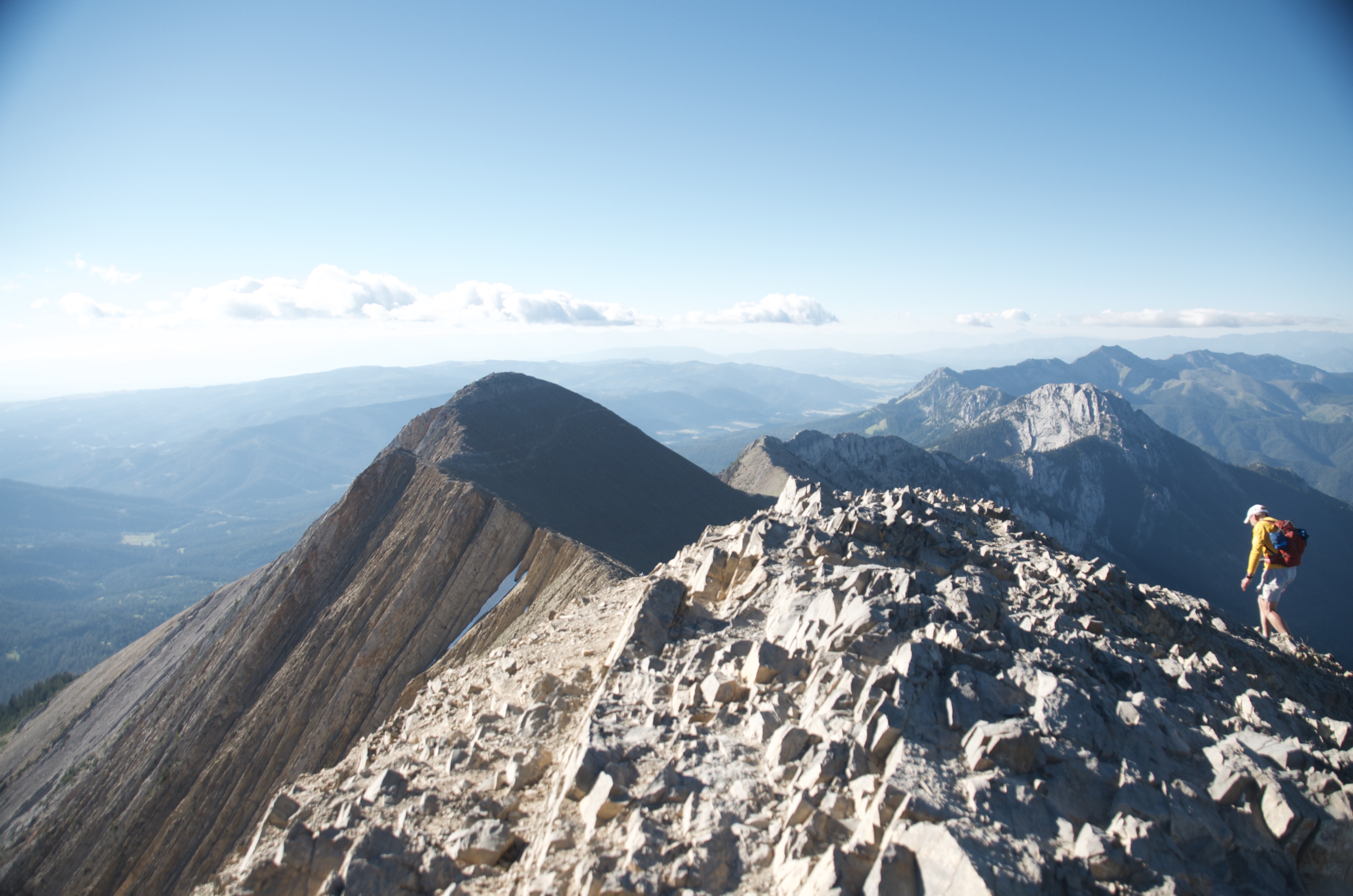 Hiker with orange pack traversing the narrow Bridger Ridge spine with a dramatic shadow peak behind and the valley far below