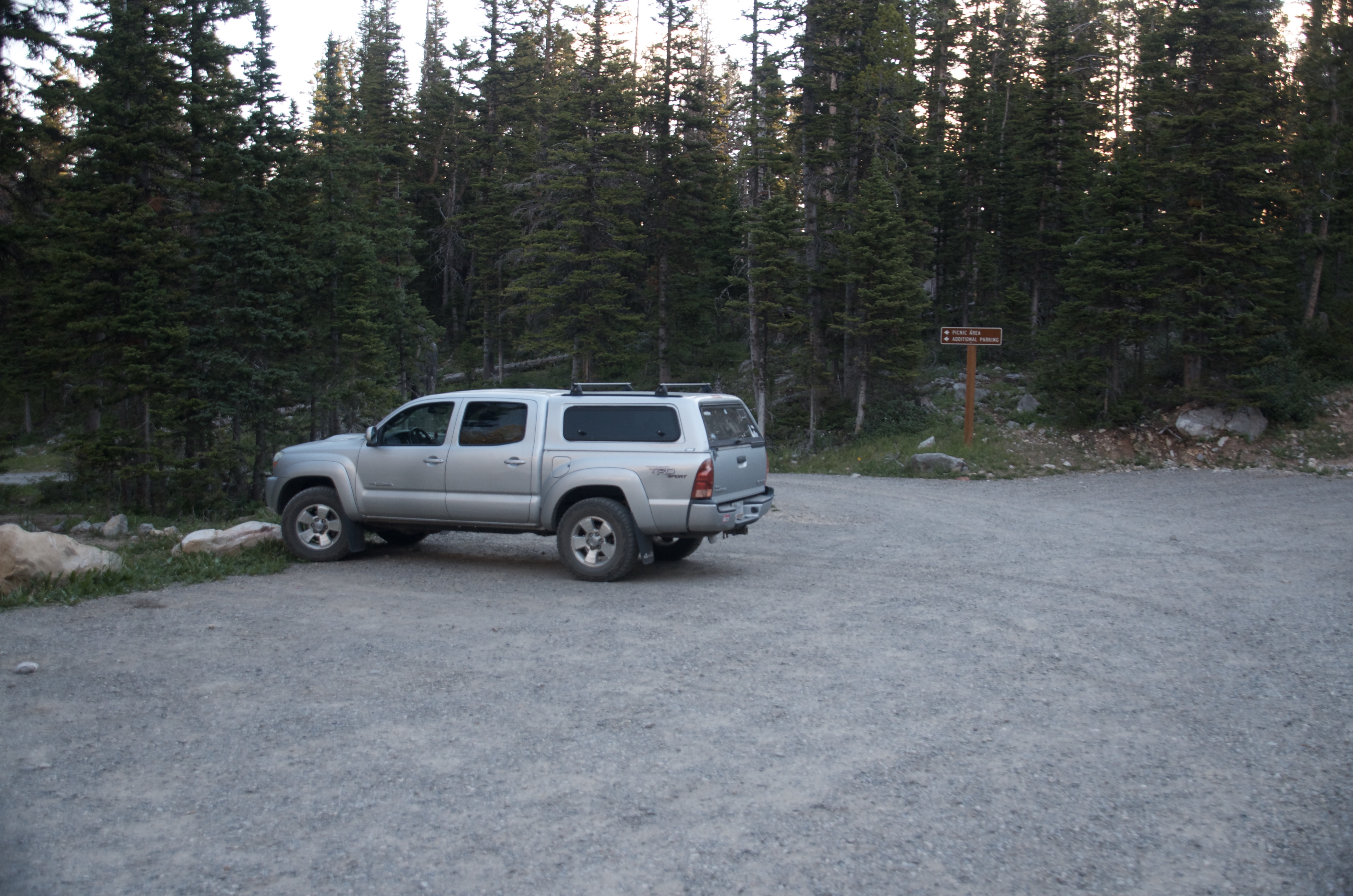 Silver truck parked at small gravel trailhead at Fairy Lake at dusk with spruce trees and trailhead sign visible