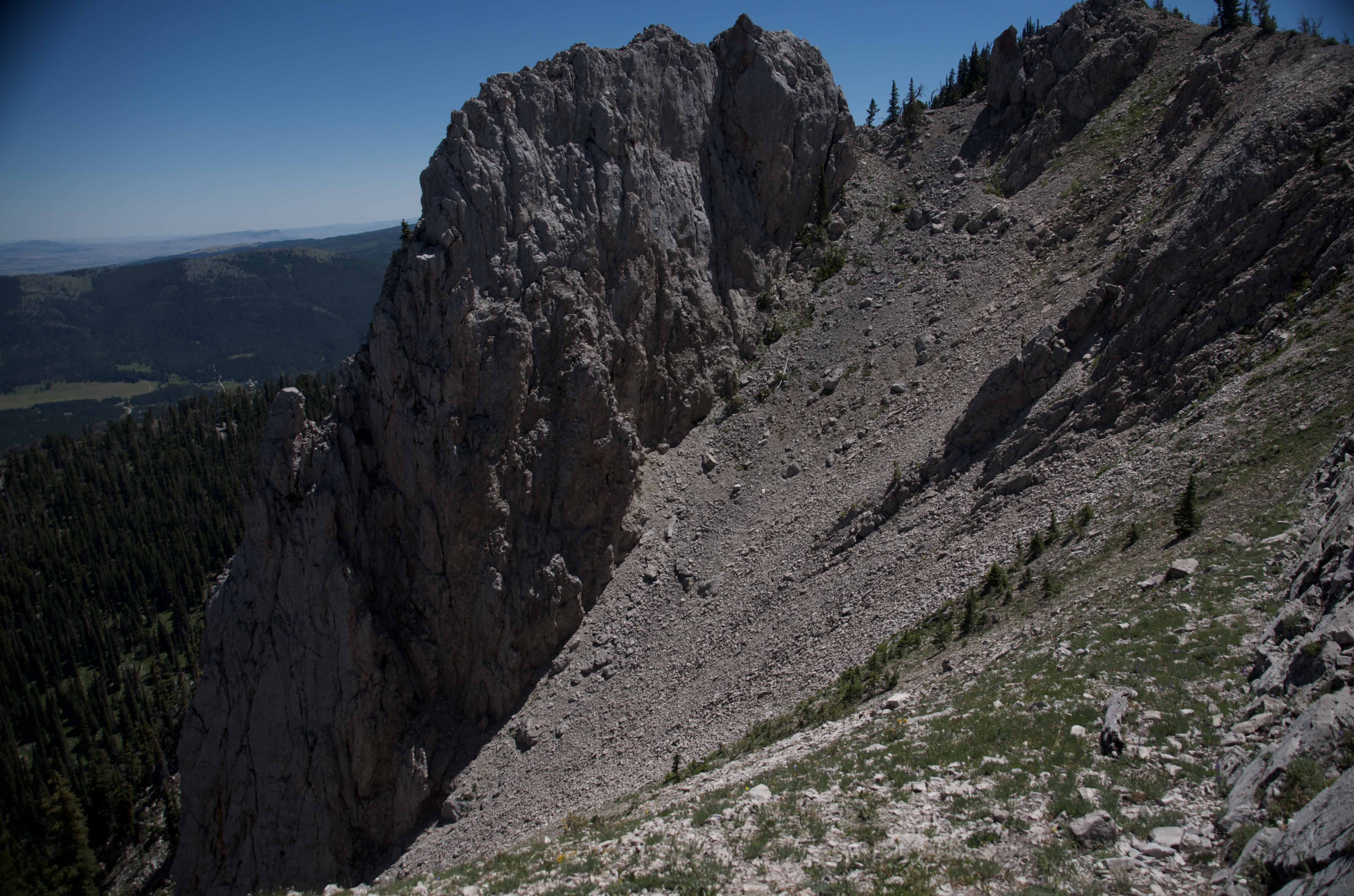 Sheer limestone cliff dropping straight off the Bridger Ridge with a hiker visible as a small figure on the talus slope far below