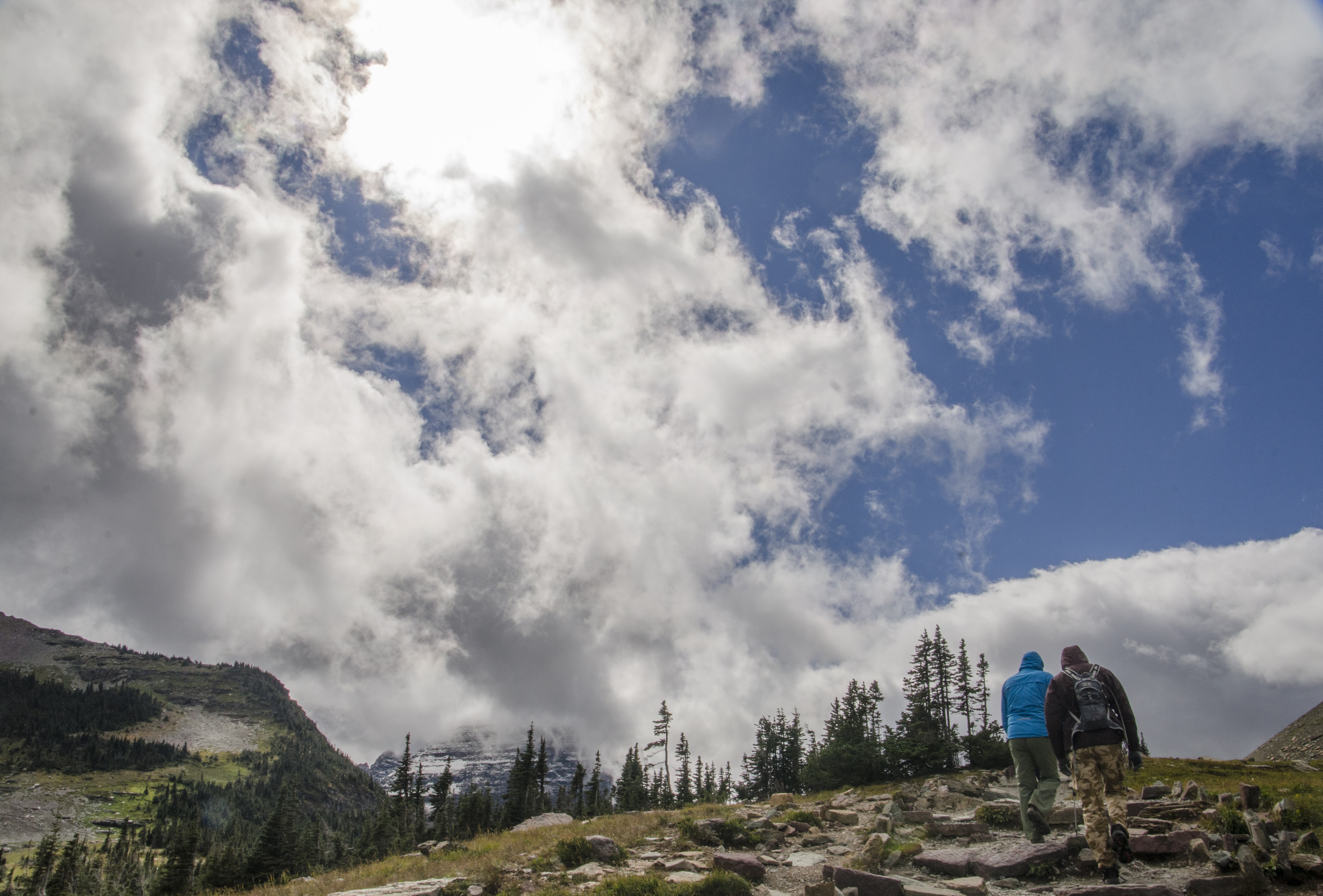 Two hikers walking on rocky trail toward mountains beneath towering clouds