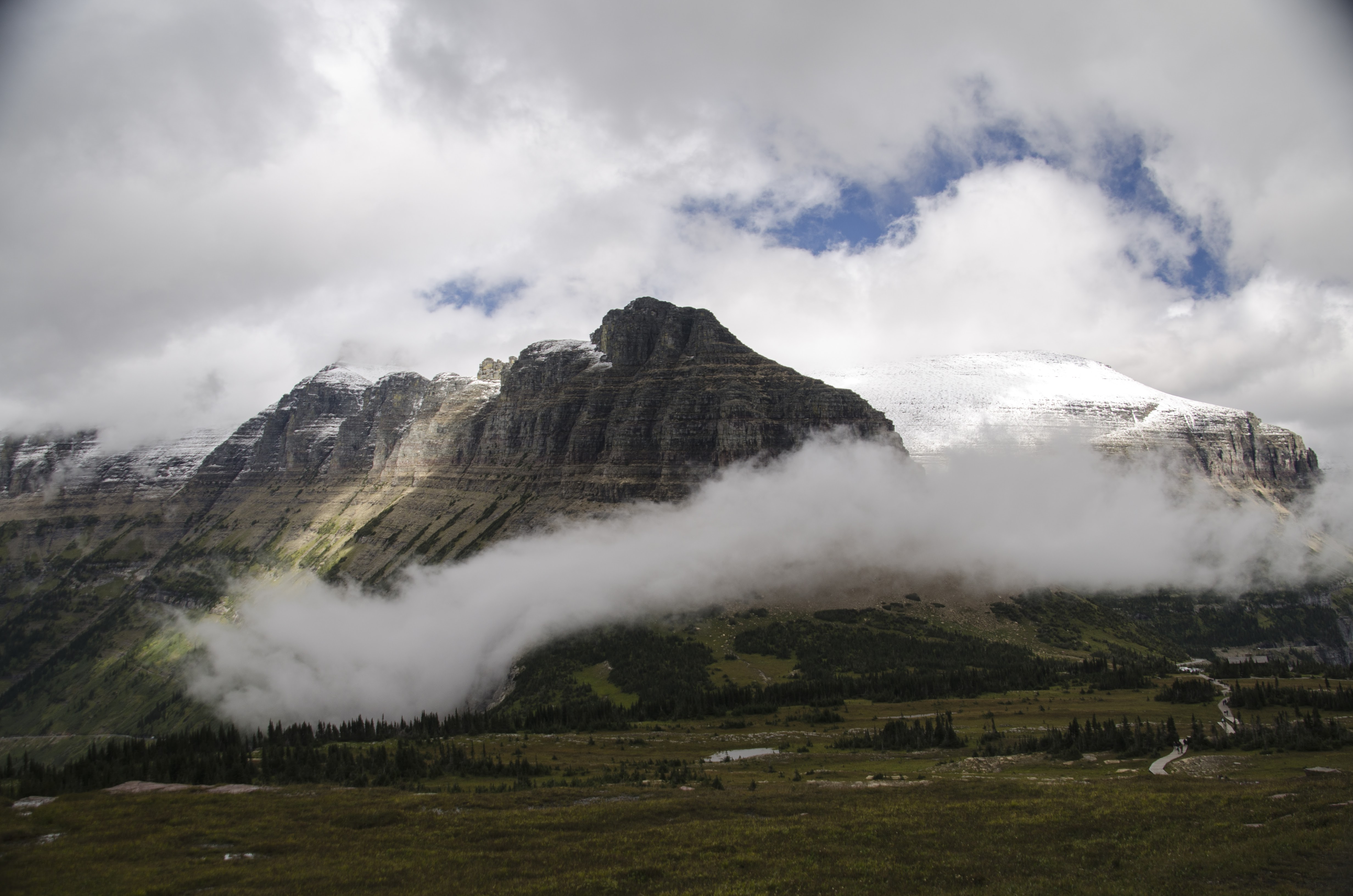 Massive snow-capped peak with clouds wrapping around the summit above alpine meadow
