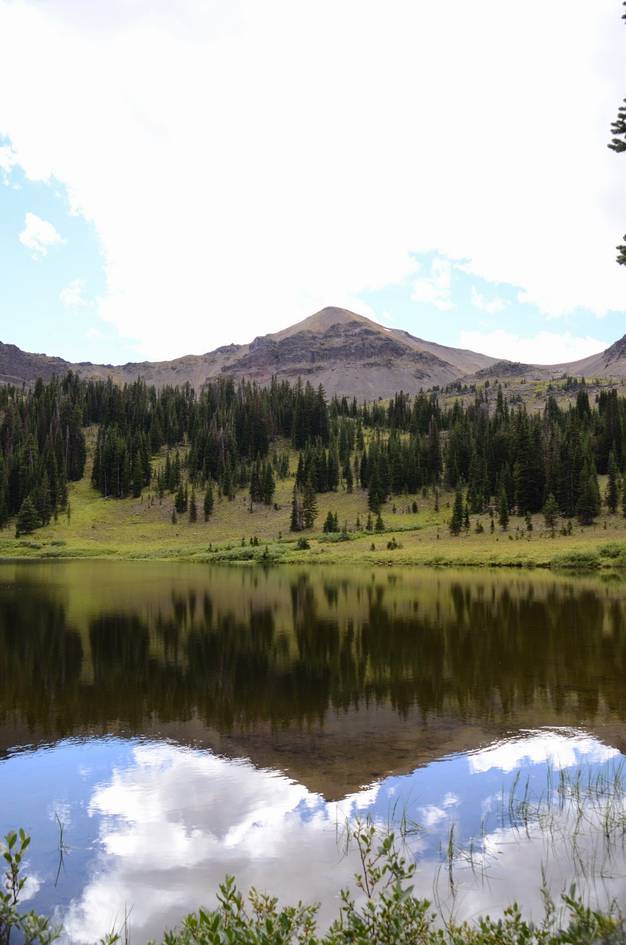Calm alpine lake reflecting Hyalite Peak with spruce forest and meadow