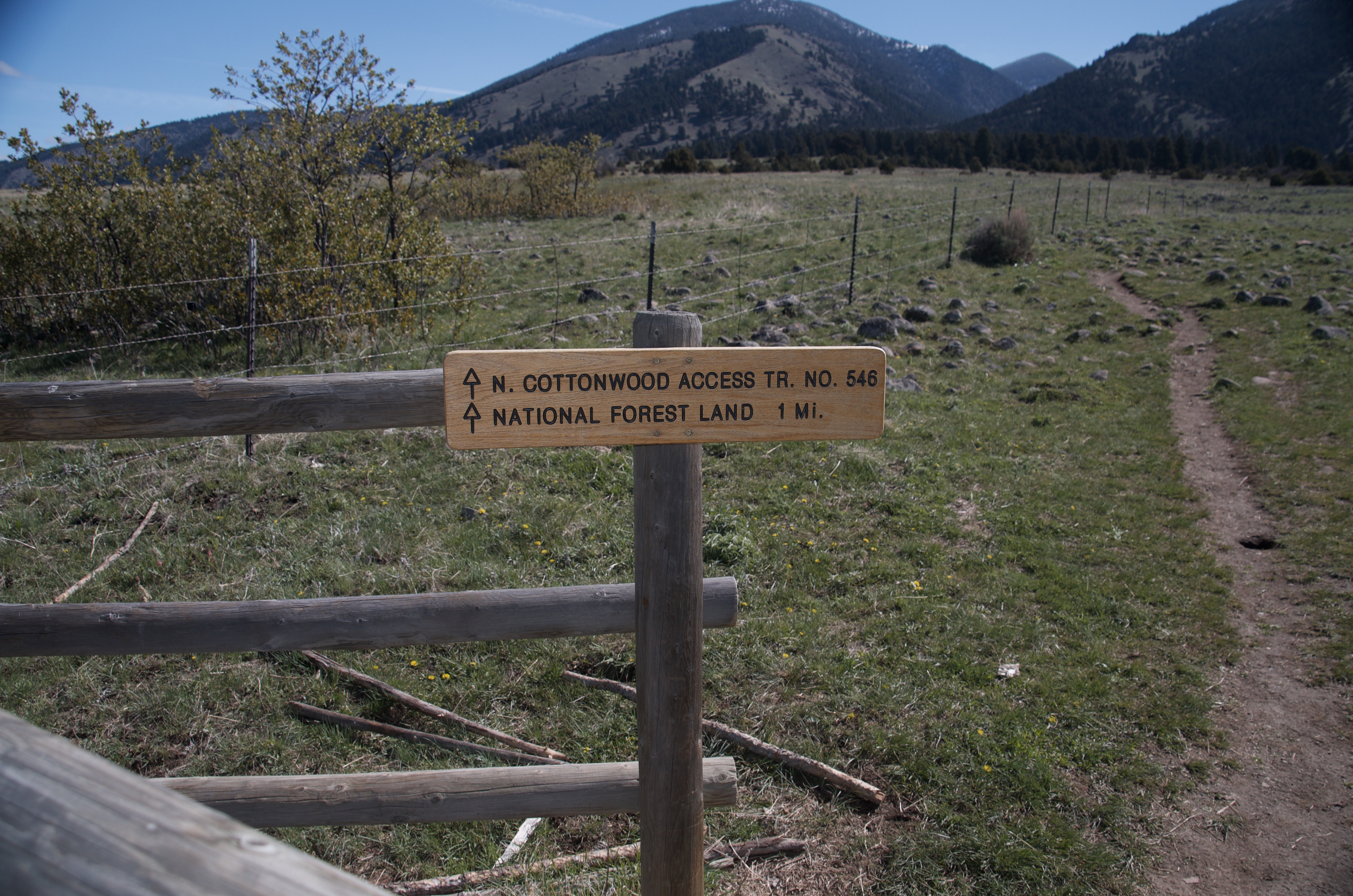 Wooden post trail sign reading N. Cottonwood Access Trail No. 548 National Forest Land 1 Mile with open ranchland behind