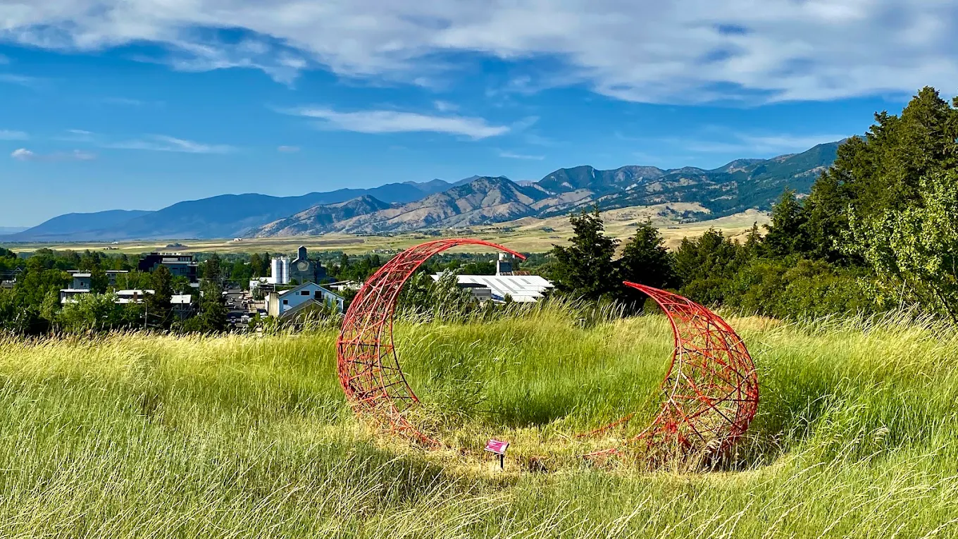 Red circular ring sculpture in green summer grass on the Burke Park hillside with Bozeman and mountains visible behind it