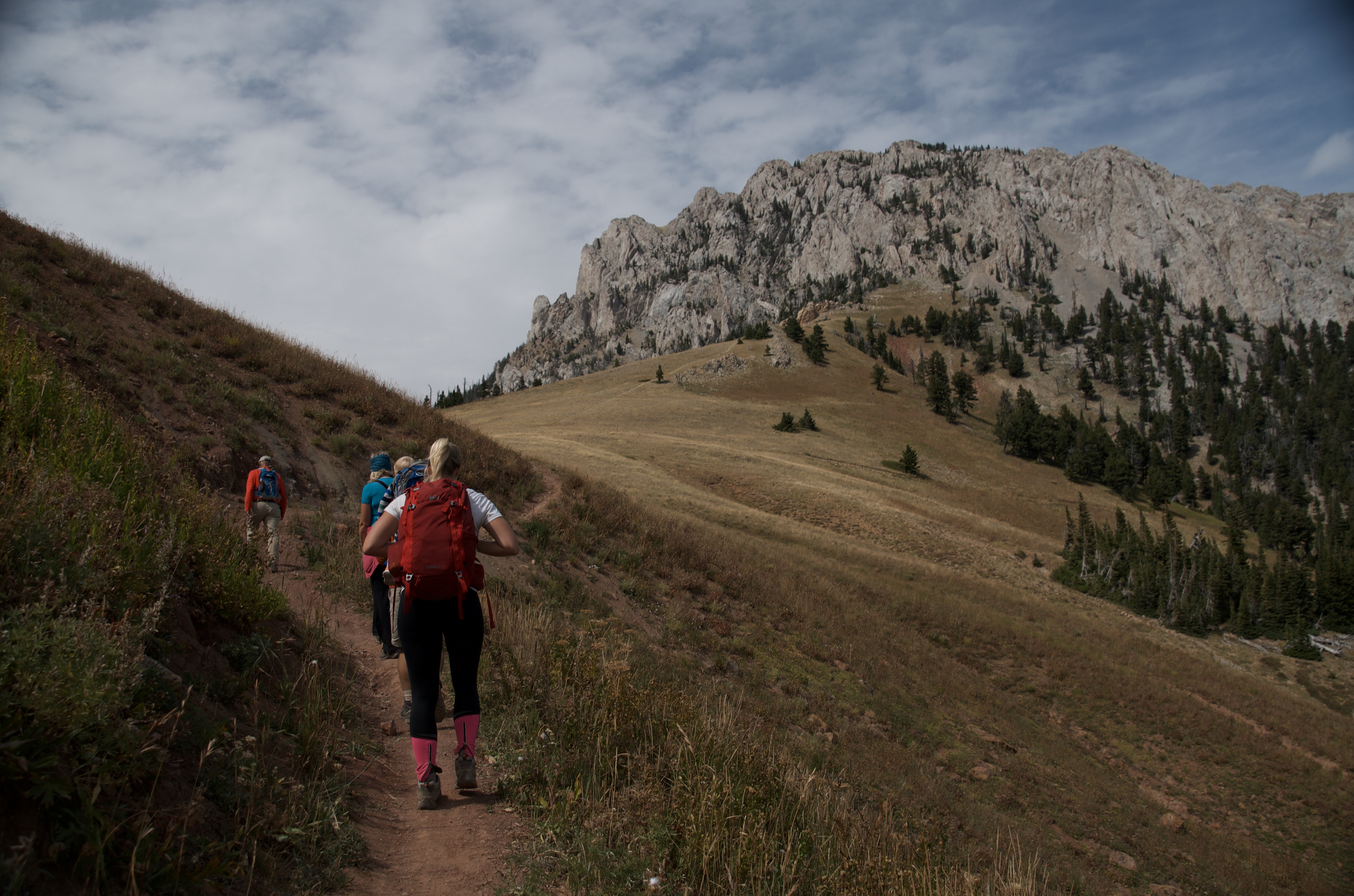 Group of hikers ascending open golden hillside with dramatic limestone cliff face of Ross Peak ahead