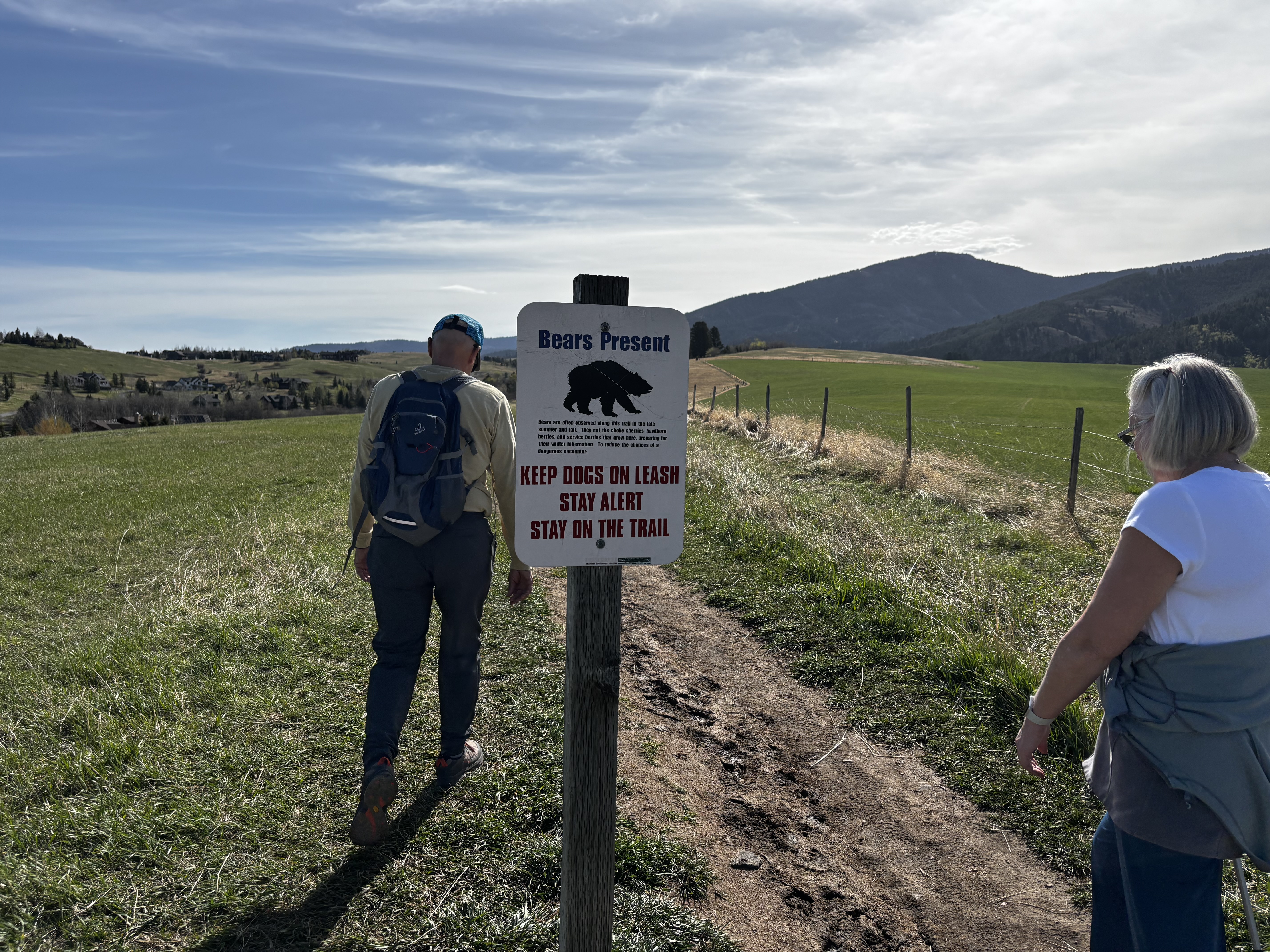 Bears Present sign reading Keep Dogs On Leash Stay Alert Stay On The Trail, with hikers and green ranch fields behind it