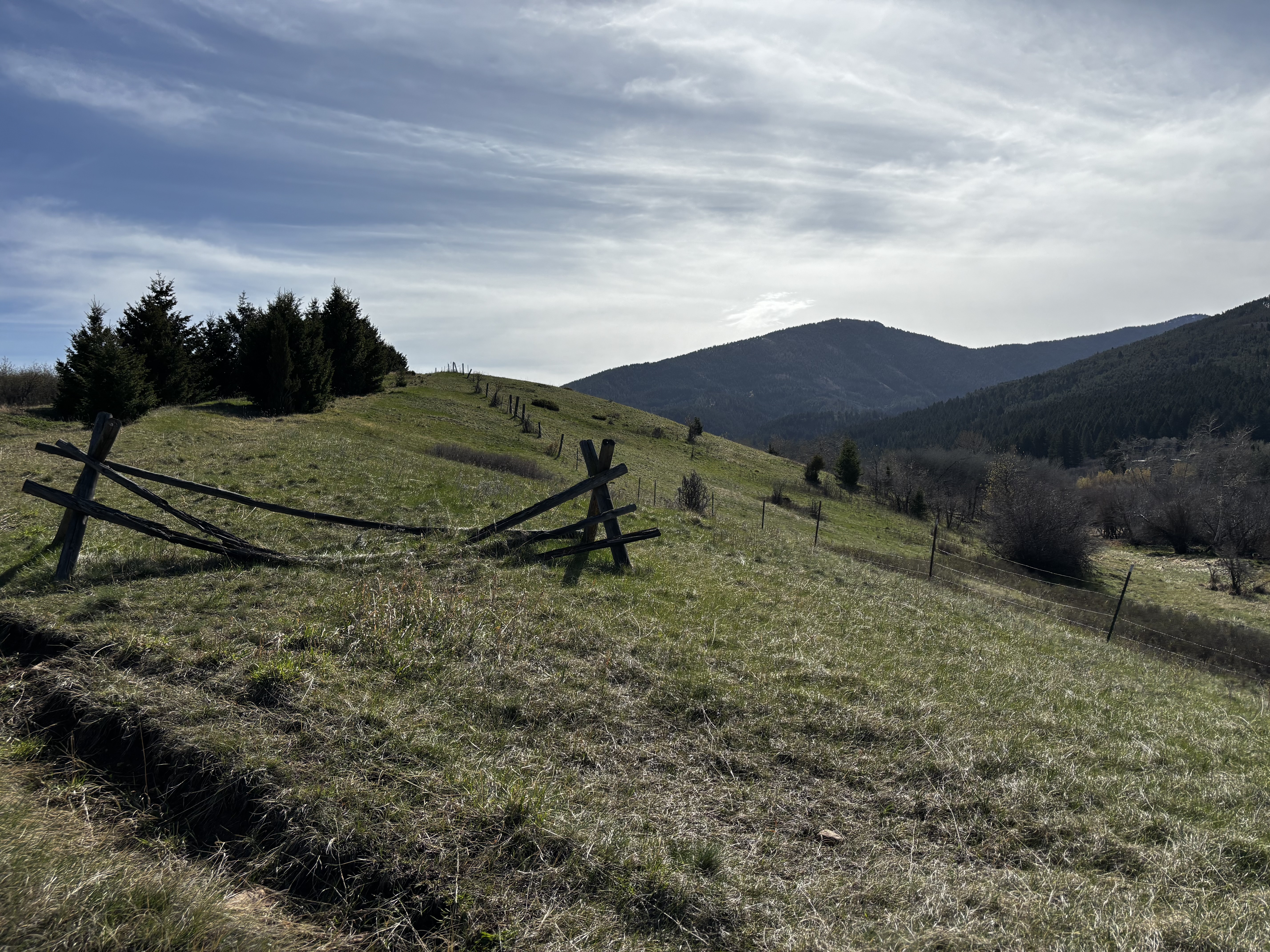 Old weathered split-rail fence leaning across a green hillside with dark forested mountains rising behind it under a partly cloudy sky