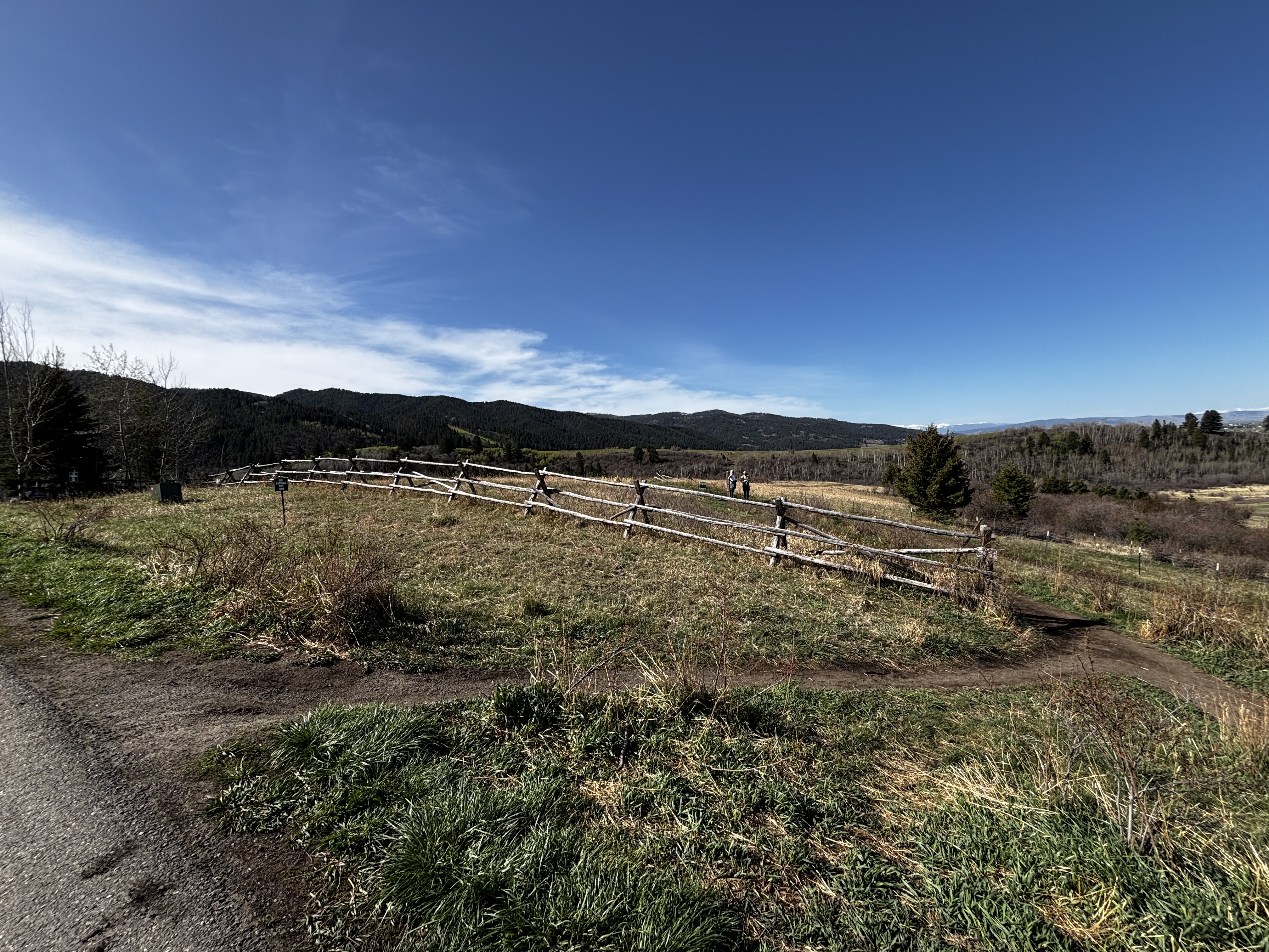 Split-rail fence running alongside the trail with rolling green hills and mountain peaks stretching across the horizon
