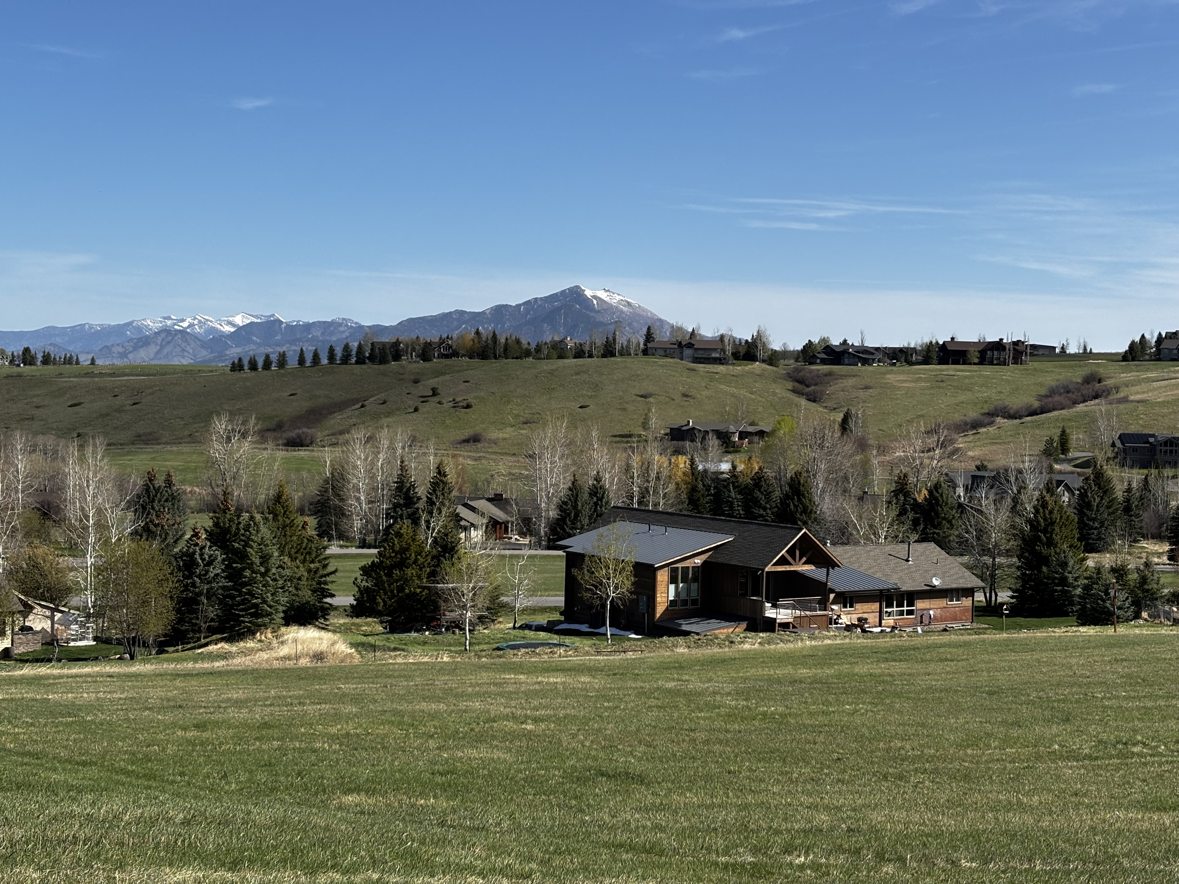 Green ranch fields and aspen trees in the foreground with a log cabin and snow-capped mountains visible across the valley