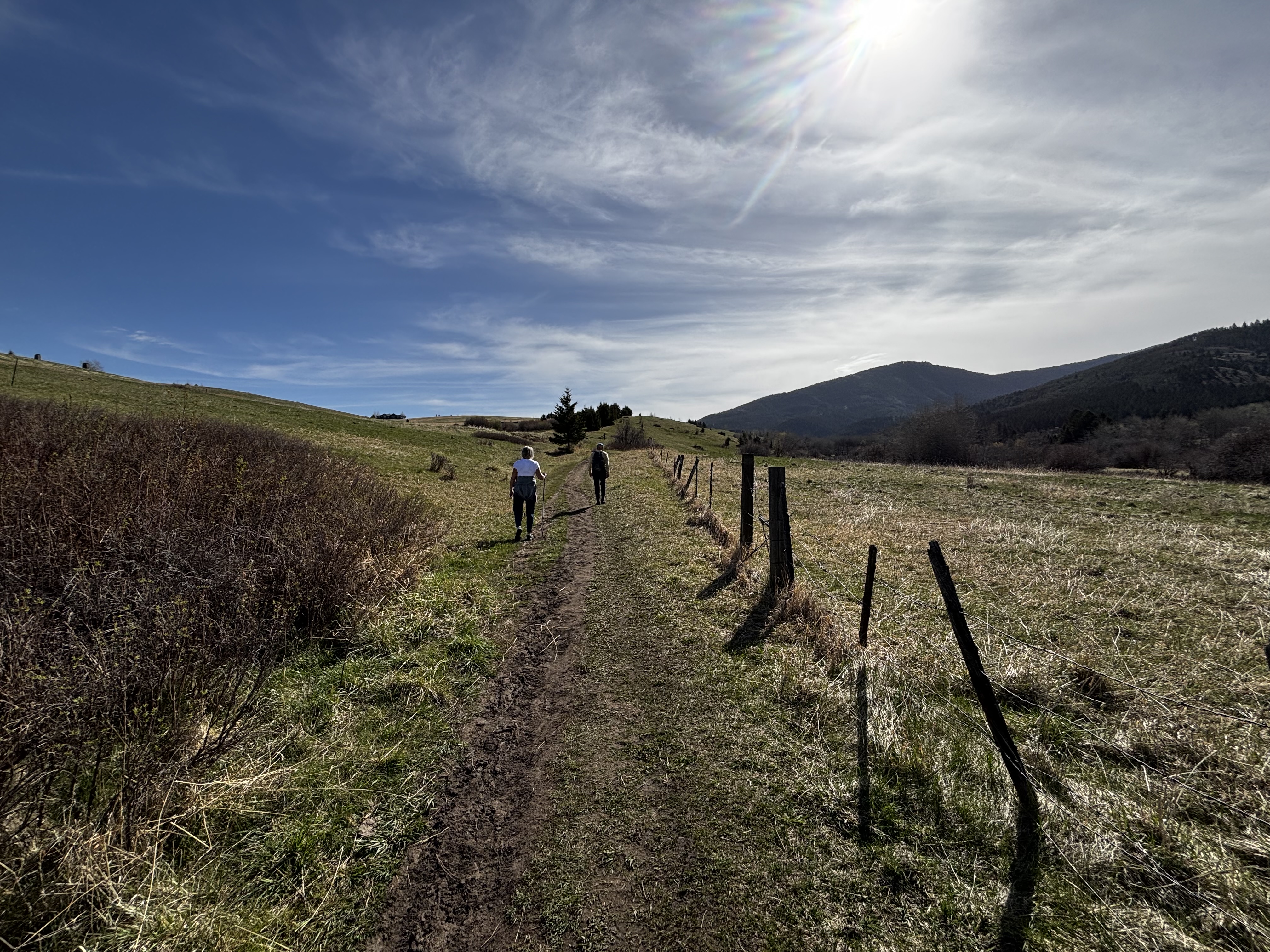 Two hikers walking a dirt trail along a fence line with mountains visible in the background