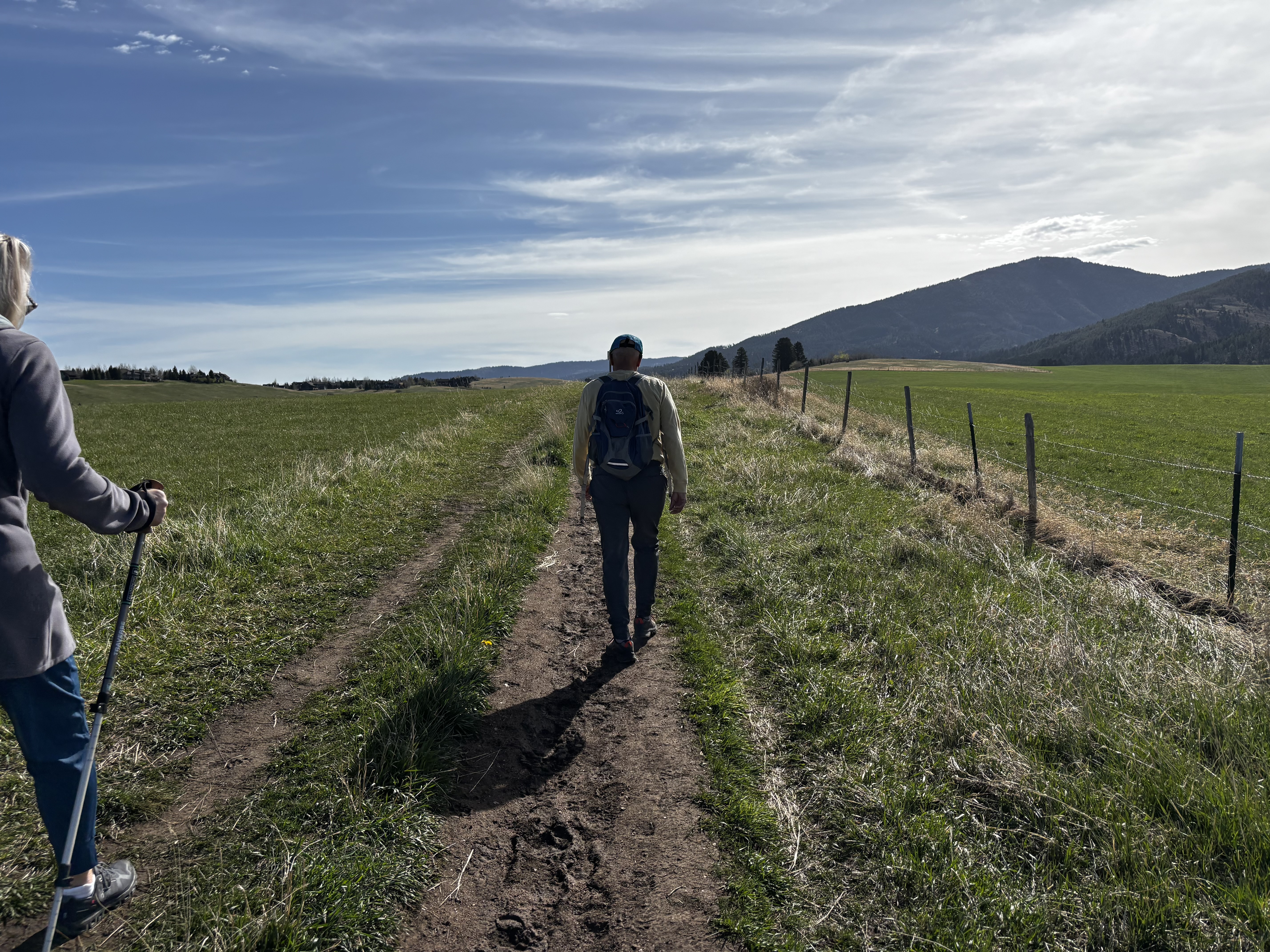 Two hikers walking a wide muddy trail through green meadows with mountains visible in the distance