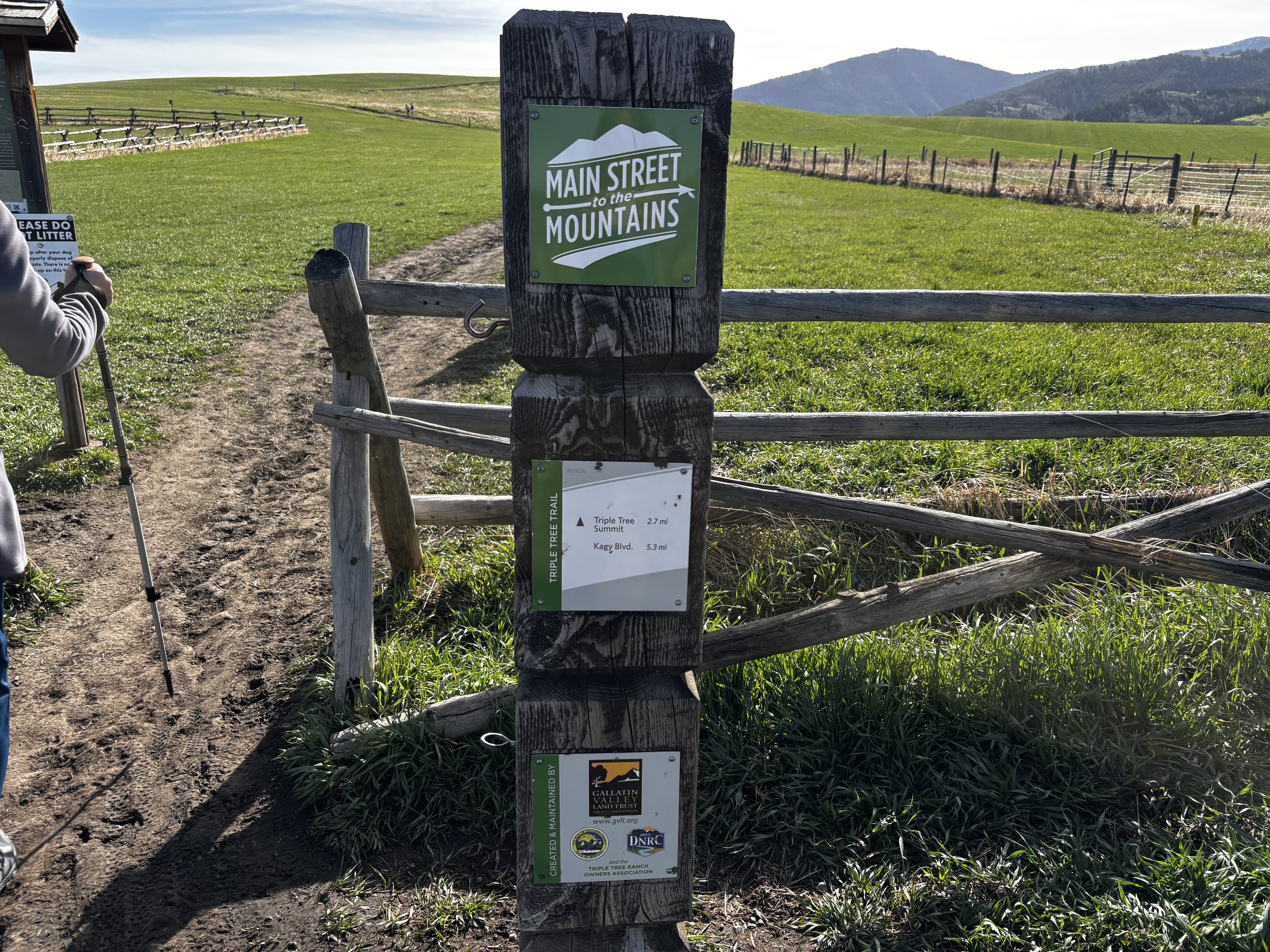 Main Street to Mountains trail sign post at the Triple Tree trailhead with green fenced ranch land behind it