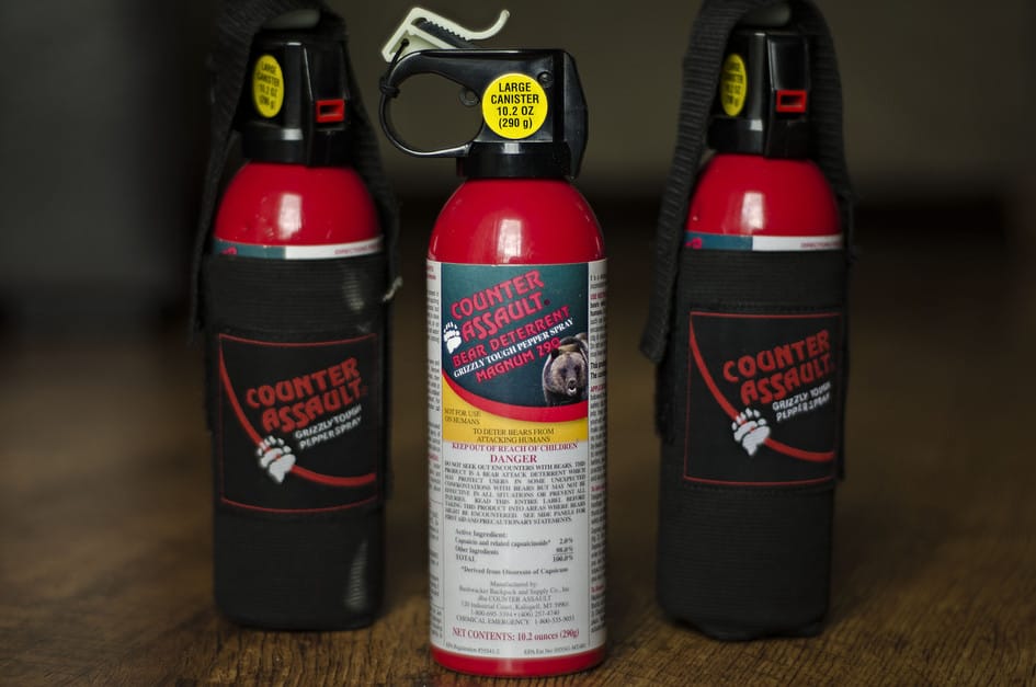 Three Counter Assault bear spray canisters with red tops and holsters on a wooden table