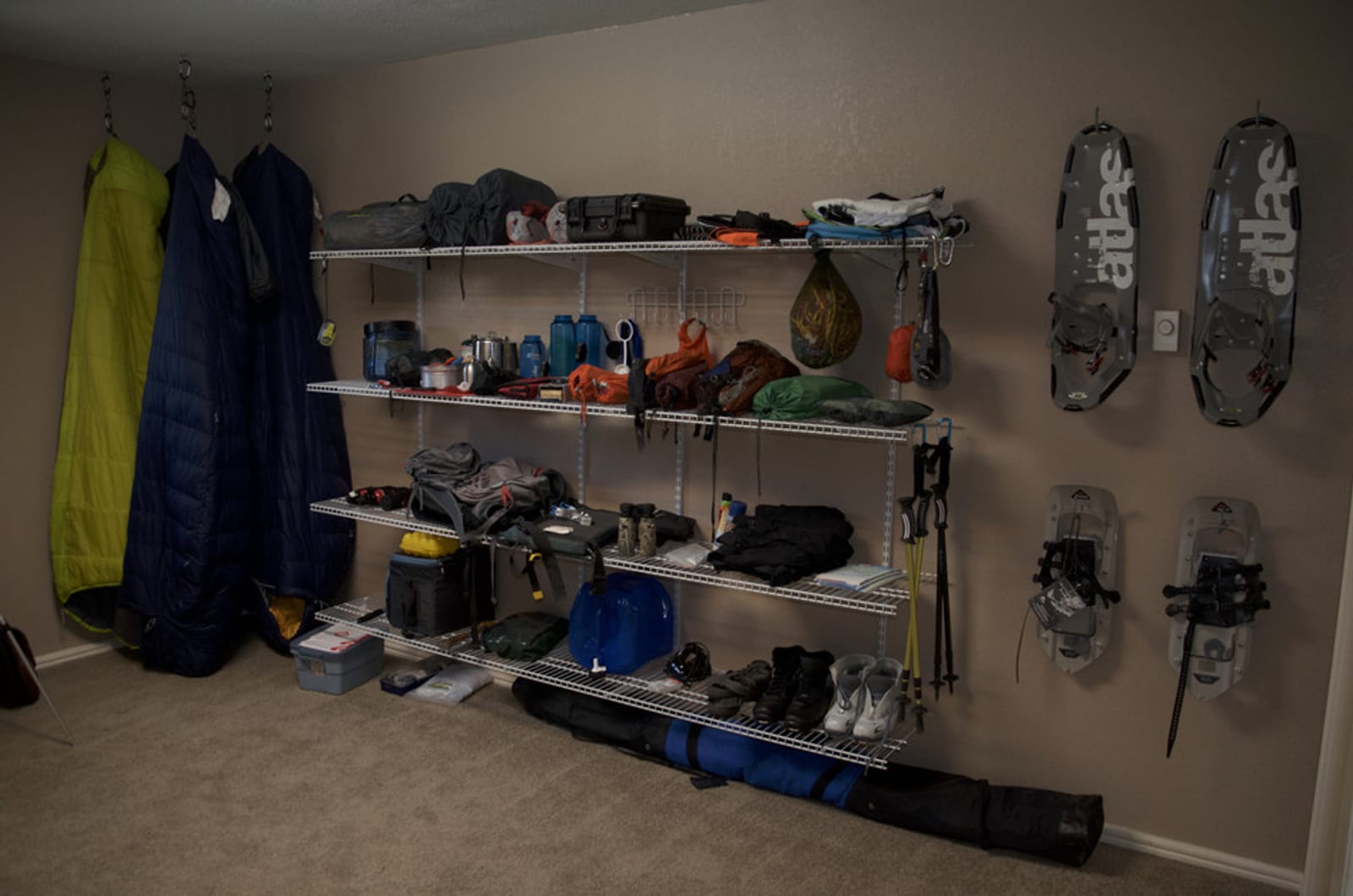 Organized gear room with wire shelving holding camping equipment, sleeping bags hanging from ceiling hooks, and snowshoes mounted on wall